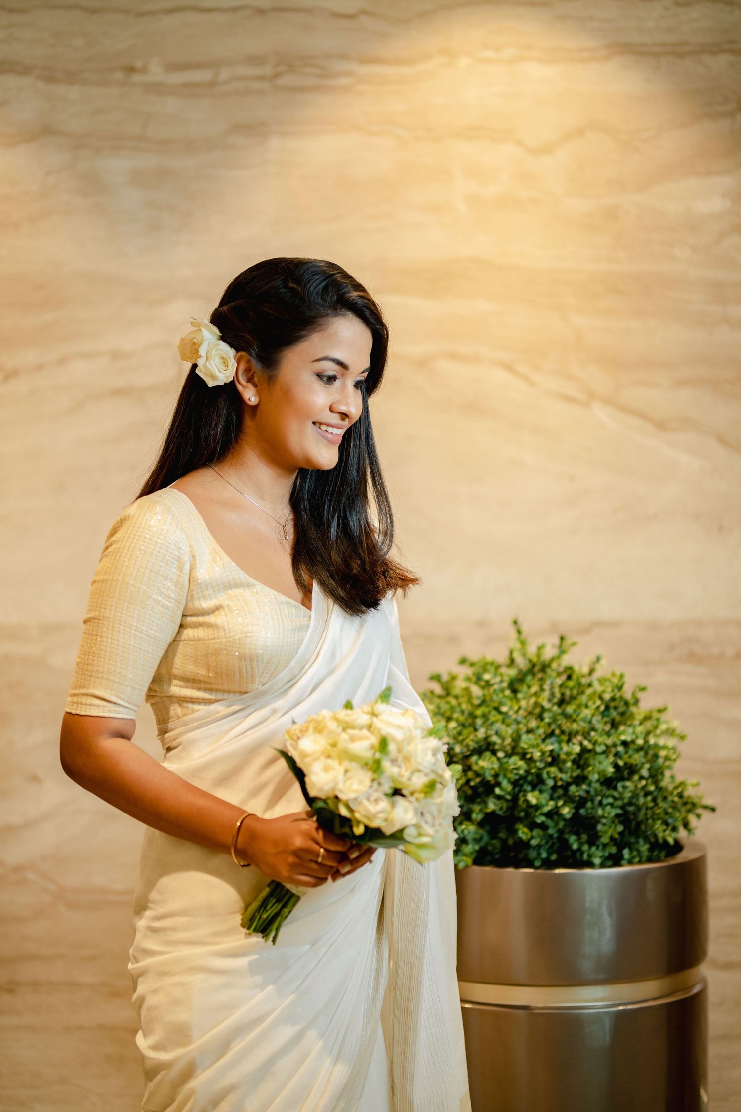A person in a white saree holds white flowers while posing near decorative plants.