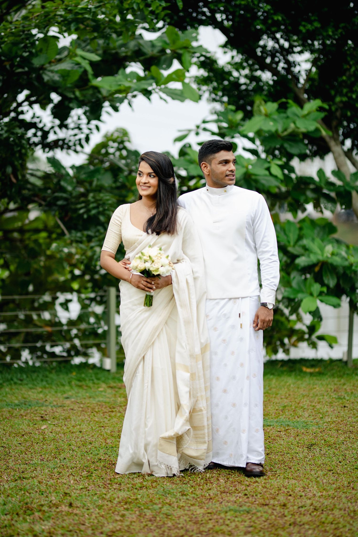 A couple in traditional white attire poses together on green grass for wedding photos.