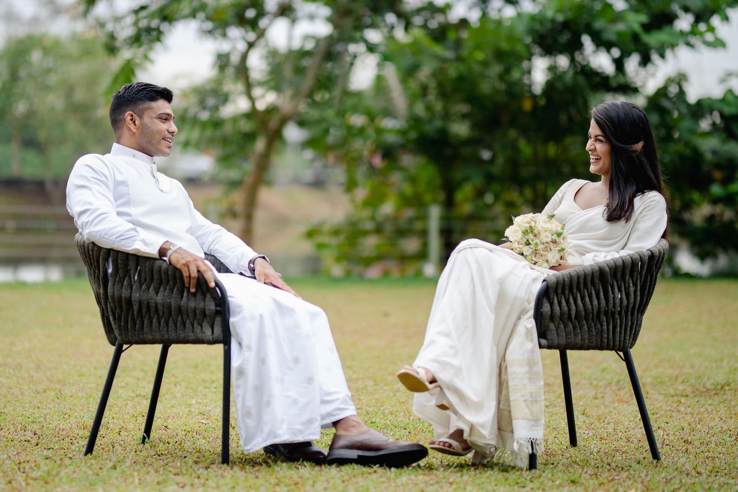 Couple in white attire sitting in wicker chairs having conversation in garden setting.