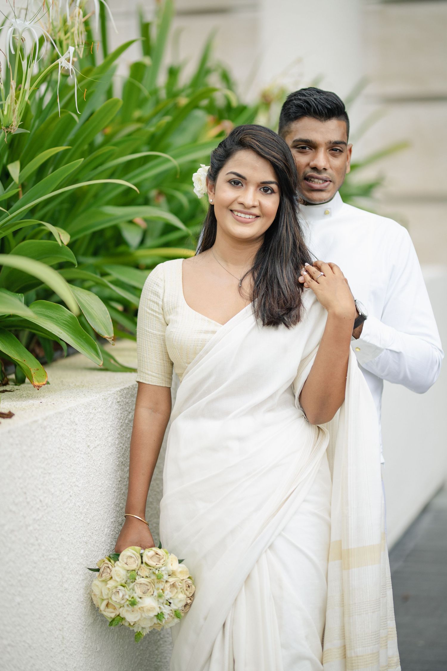A couple in traditional white attire pose together for romantic engagement photos with lush greenery in the background.