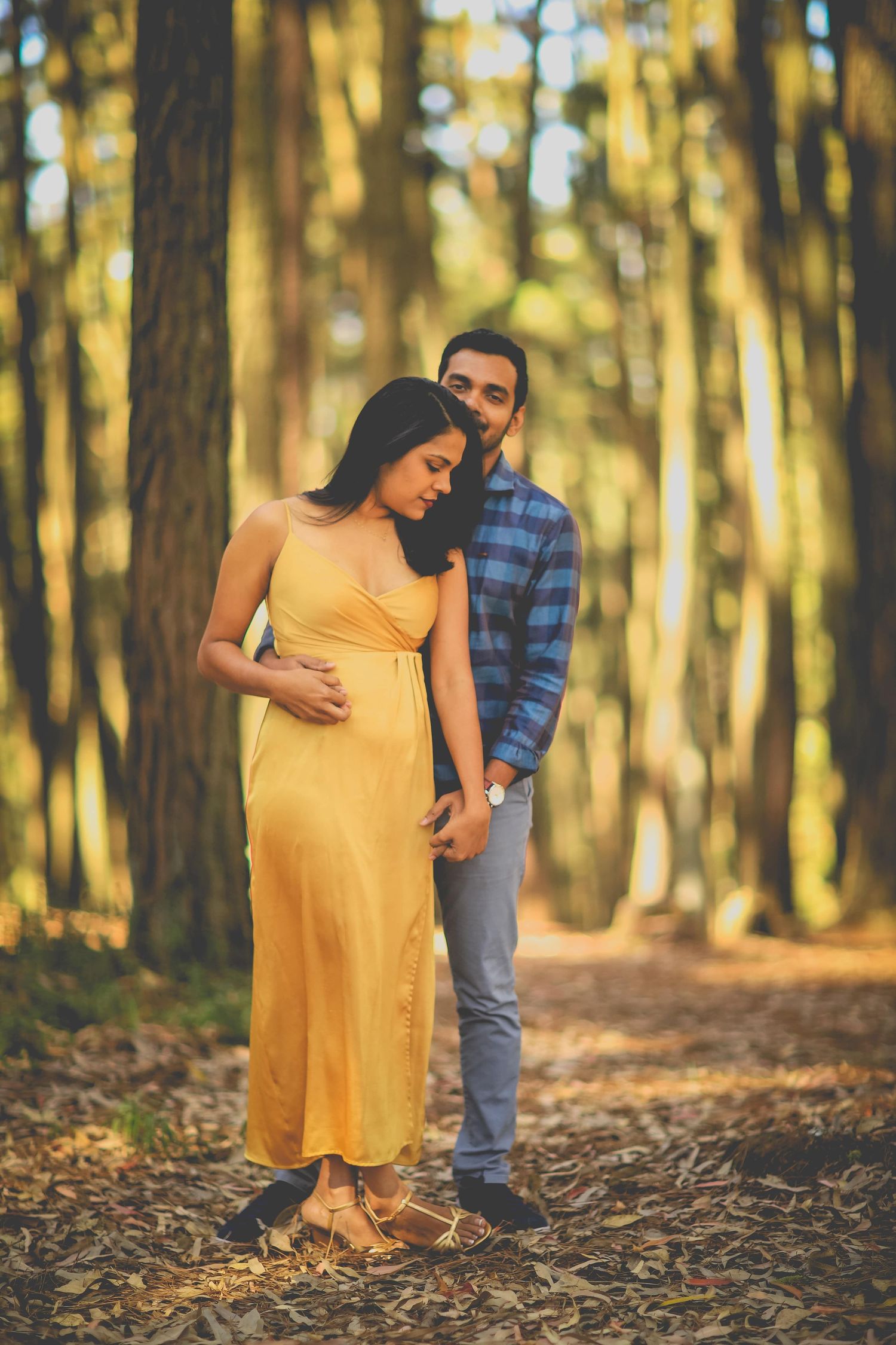 A couple shares a tender moment during their maternity photoshoot in a golden-lit forest with fallen leaves.