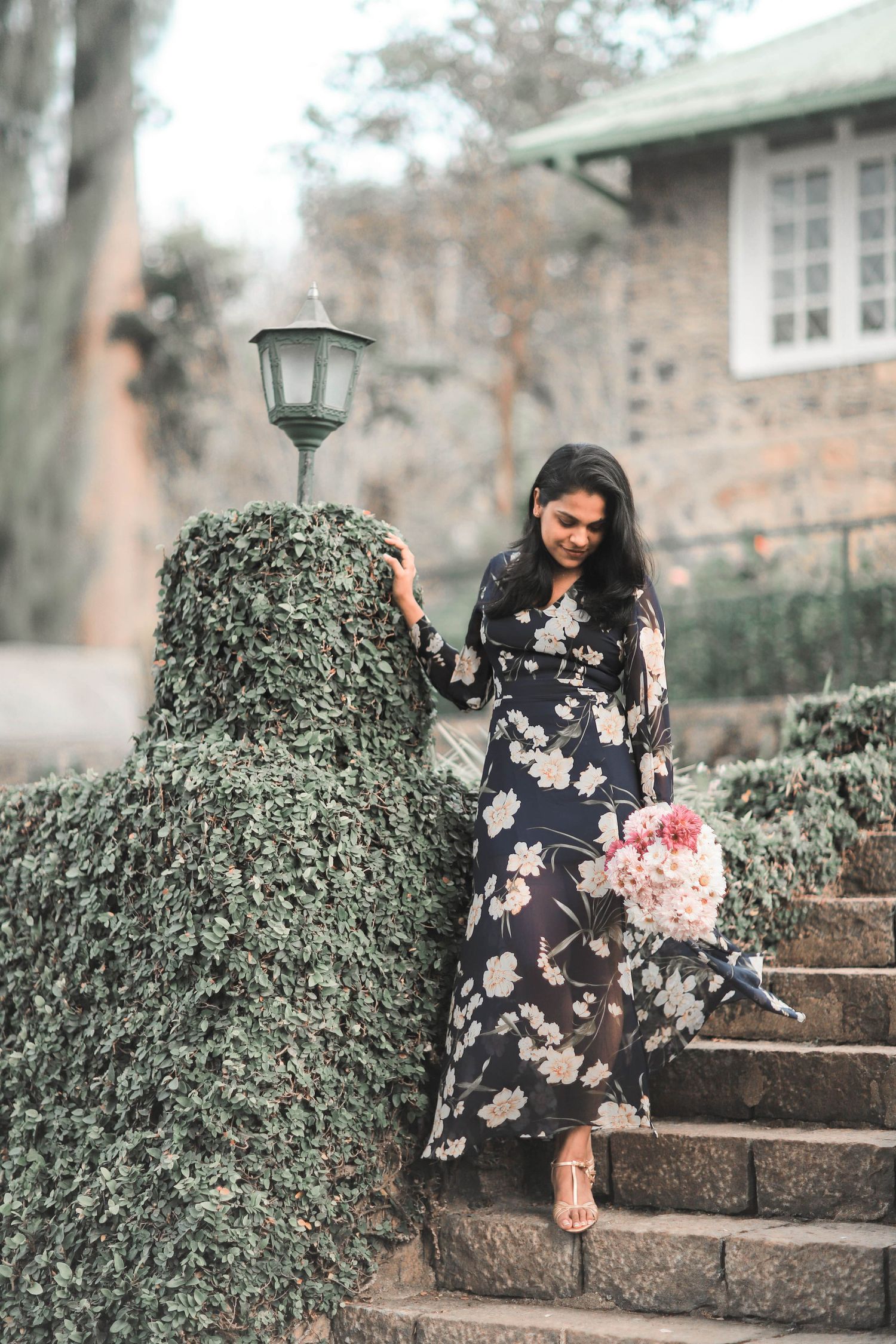 A woman in a floral black dress sits gracefully on stone steps surrounded by ivy-covered walls.