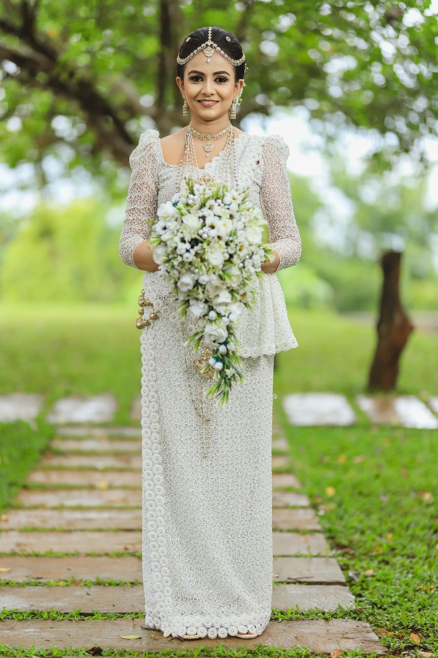 Bride in elegant white lace dress holding cascading white bouquet on garden pathway.
