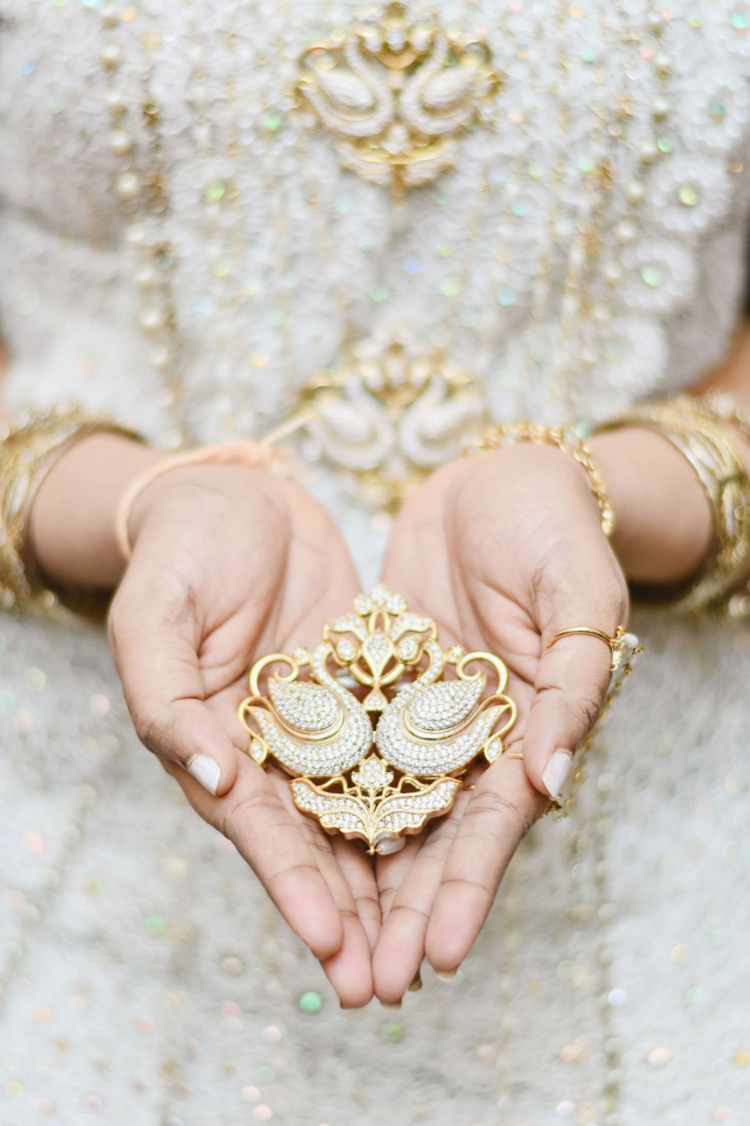 Hands holding an ornate gold and pearl brooch against white lace fabric with matching embroidered details.