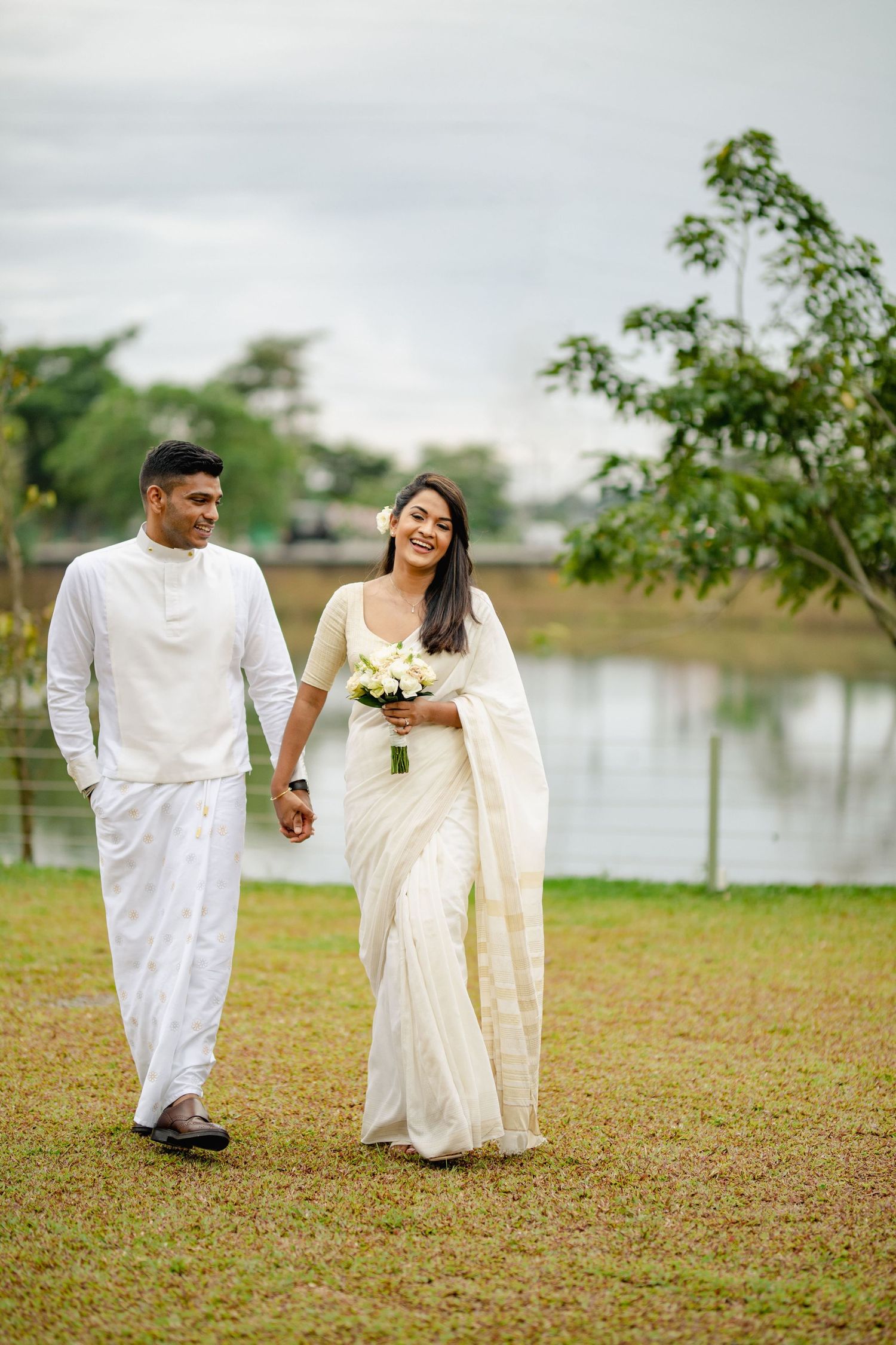 A couple in traditional white attire walks together by a lake, with the bride holding a bouquet of flowers.
