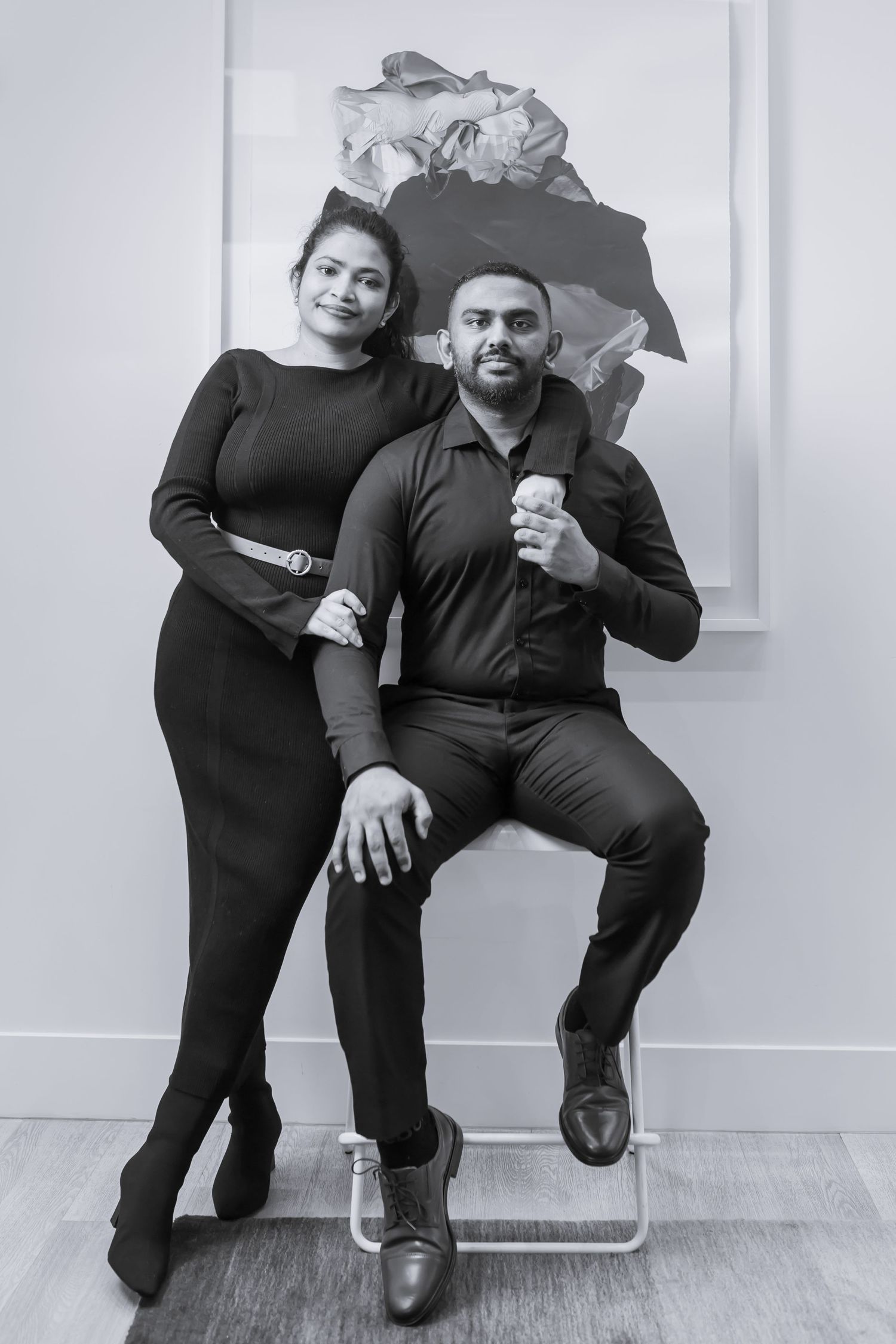 Black and white portrait of a couple in matching dark outfits posing together against a light wall with artwork.