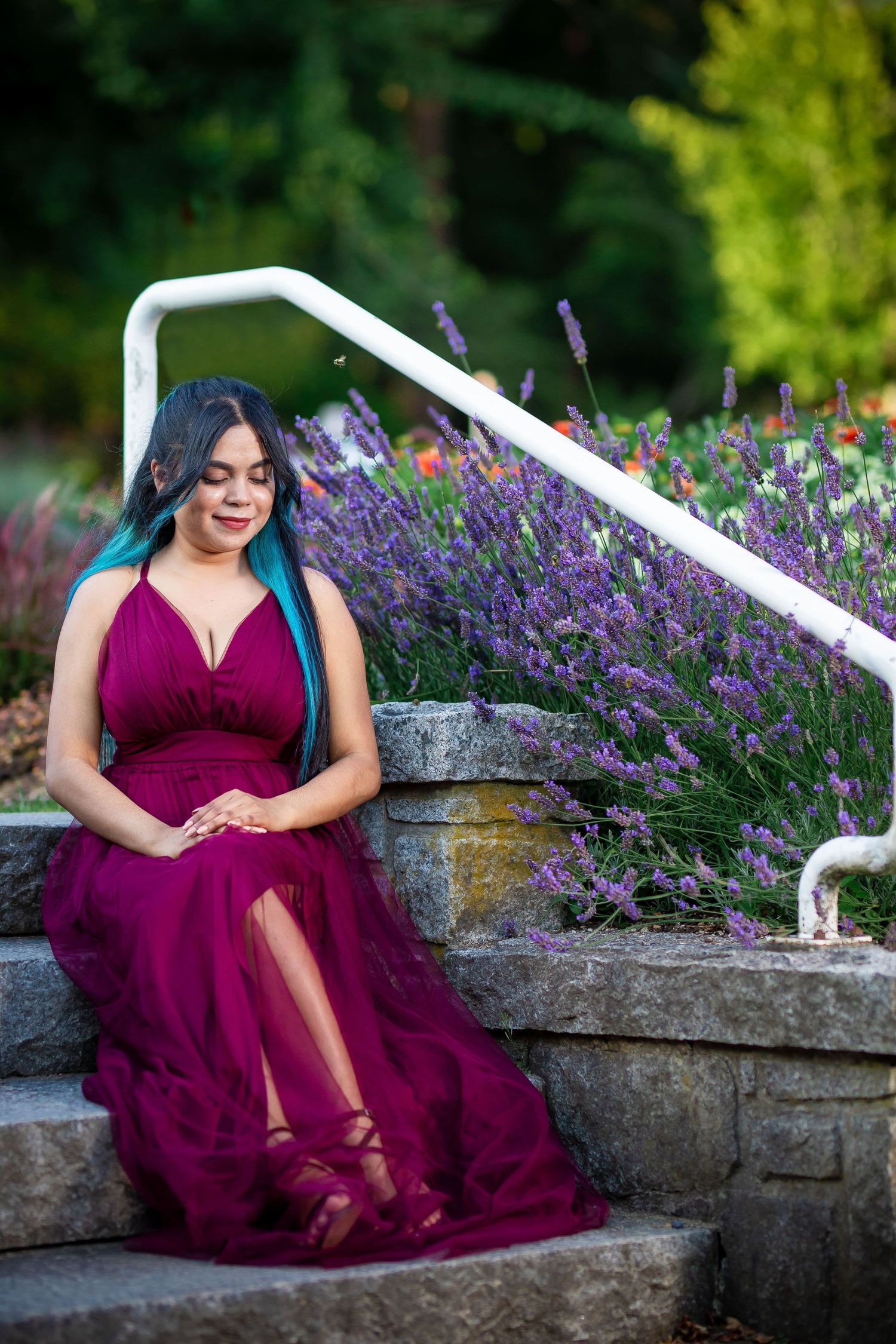 A person in a burgundy evening gown poses gracefully near purple lavender flowers and a stone wall in a garden setting.