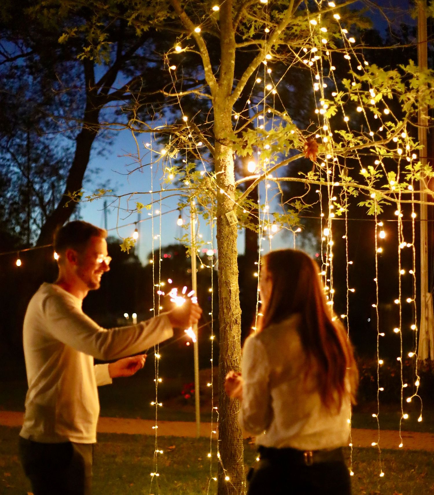 two people talking outdoors in the evening at an event, under a tree filled with fairy lights