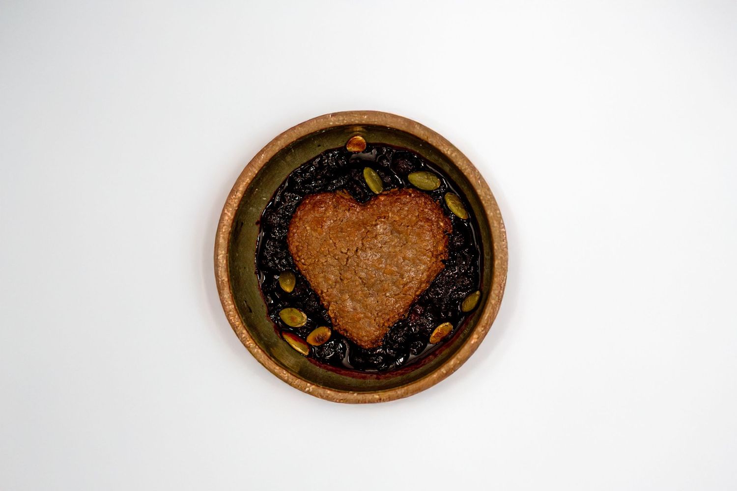 Heart-shaped cookie nestled in a rustic bowl with huckleberry compote and pumpkin seeds, viewed from above on a white surface.