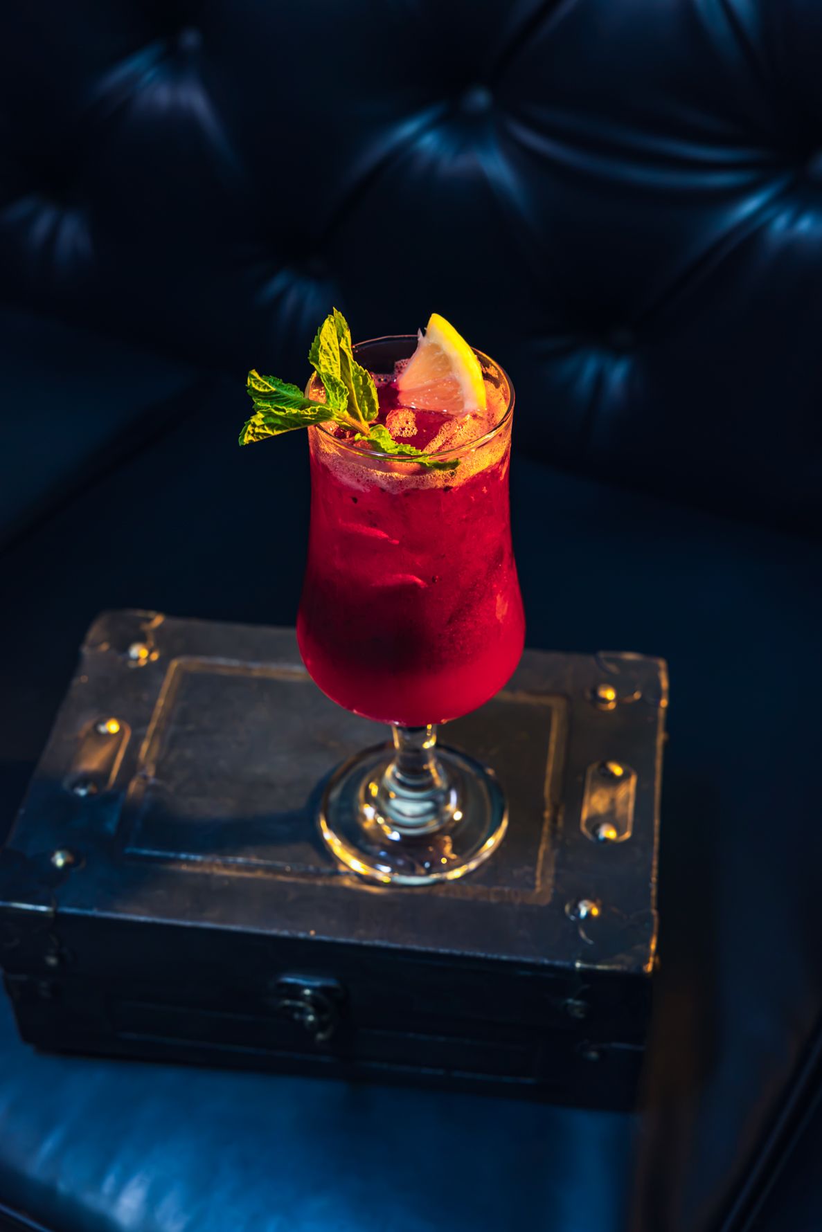 Red cocktail garnished with mint and lemon slice, served in a glass on a vintage case beside dark leather sofa. B. Steele Images