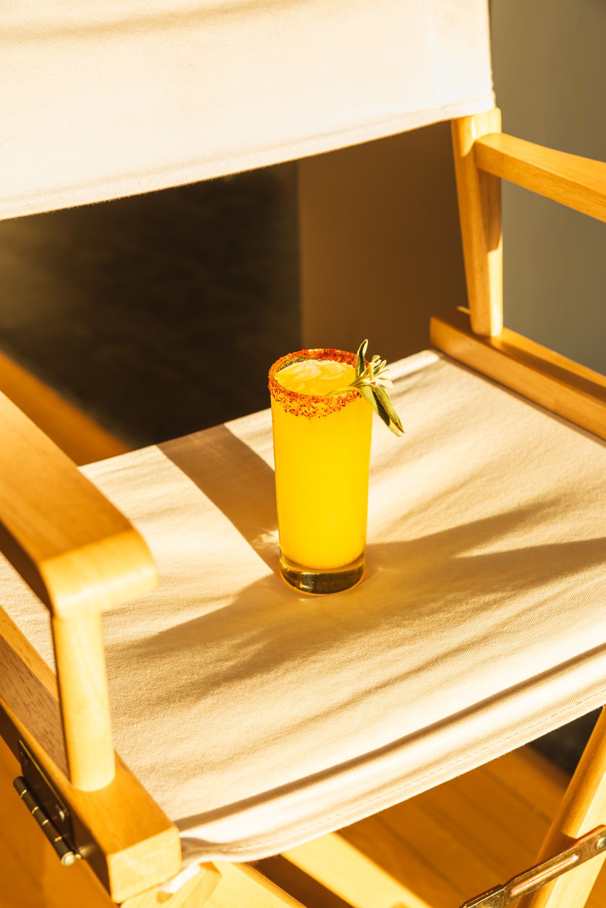 A tall yellow mocktail with a salted rim and sage garnish sits on a wooden director's chair in warm sunlight. B. Steele Images