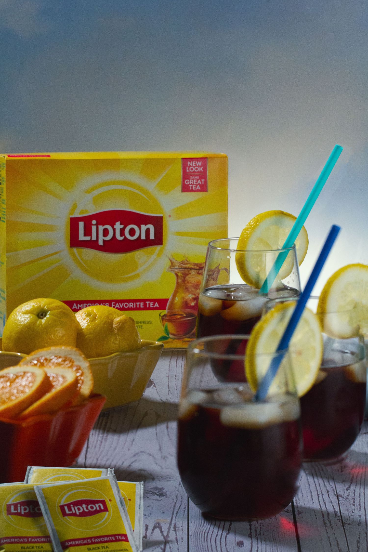 A tablescape of Lipton iced tea drinks garnished with lemon slices alongside a yellow Lipton tea box on a light grey wooden table.