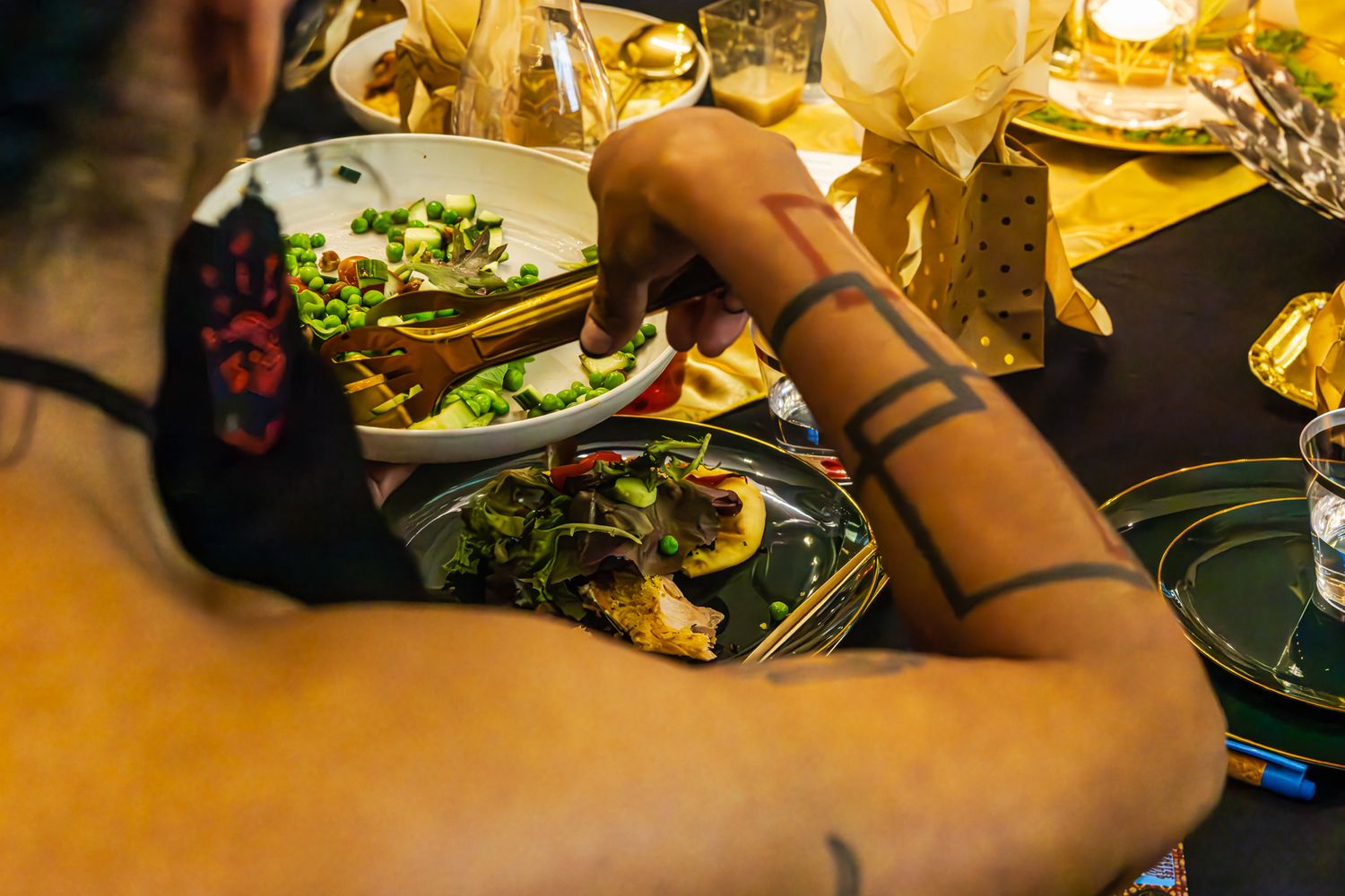 Person with tattoos serving food at a dinner table with colorful dishes and golden decorations. B. Steele Images. Food & Beverage photograph