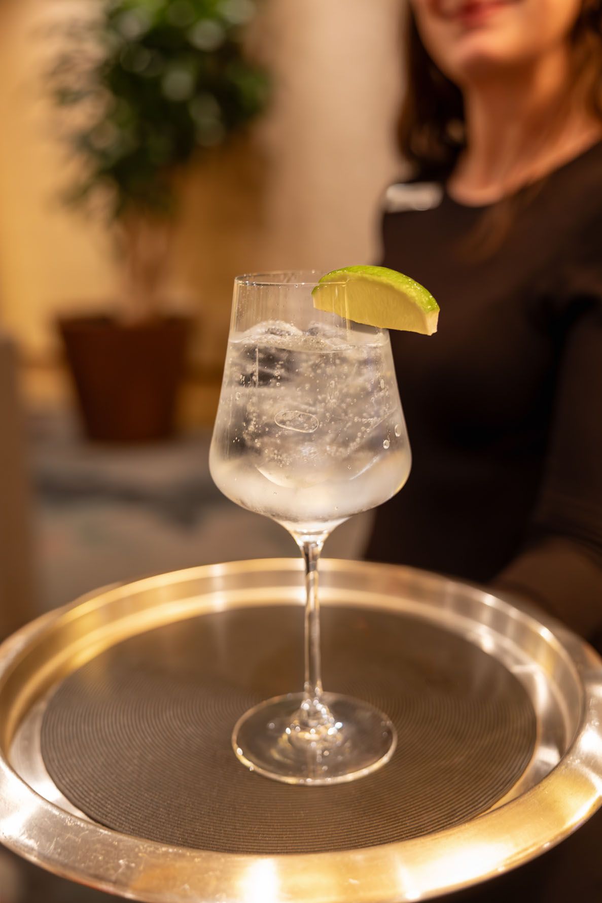 A mocktail glass filled with a clear beverage and garnished with a lime wedge sits on a metallic serving tray.