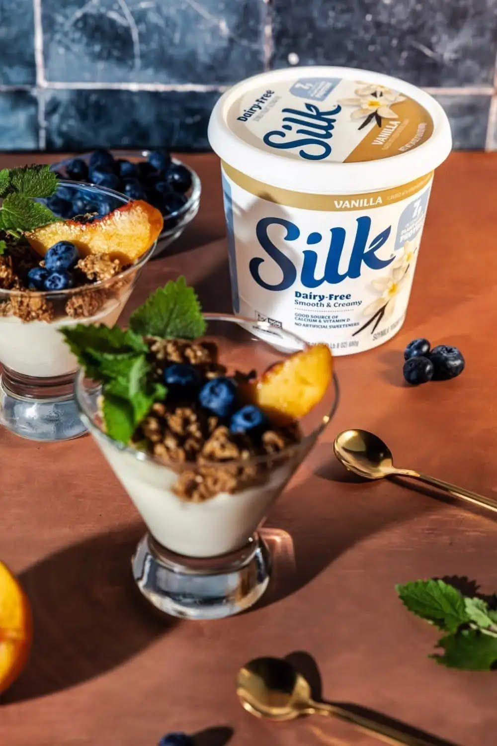 A series of yogurt parfaits with blueberries, granola, and mint garnish arranged on a wooden surface with Silk brand containers.