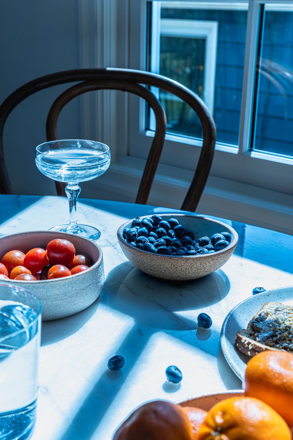 Blue-toned table scene with bowls of blueberries and cherry tomatoes, a coupe glass, and fresh fruit by a sunlit window.