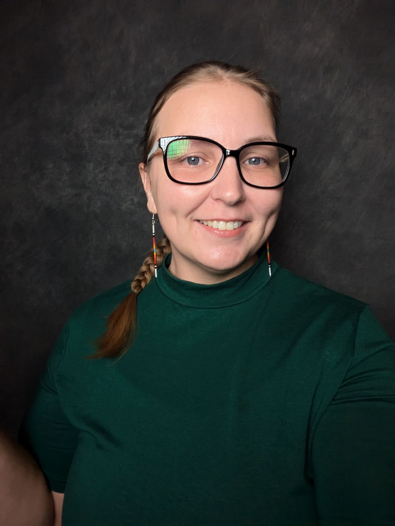 Professional headshot of Brigit against dark background showing her in a green turtleneck, glasses, and side braid with beaded earrings.