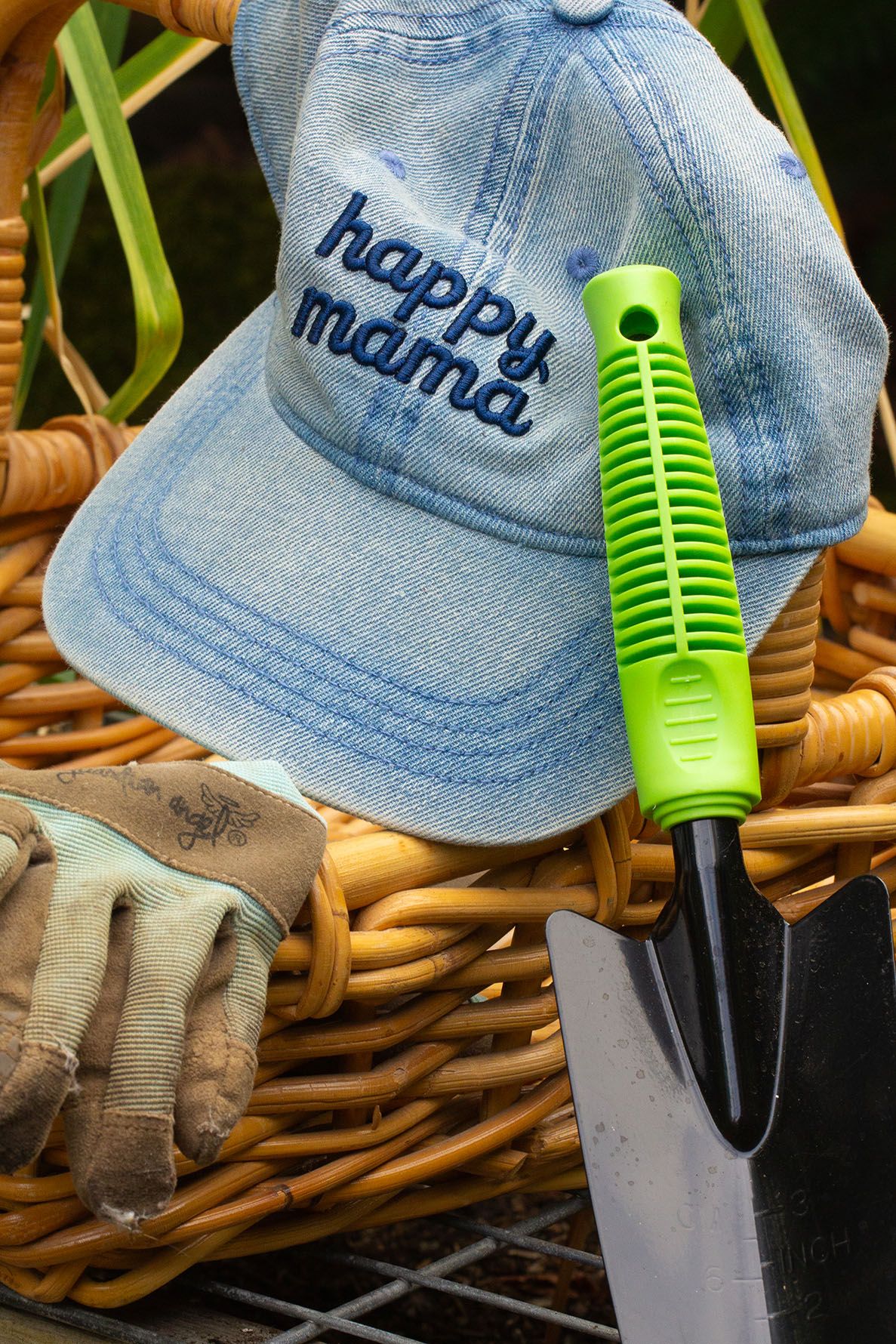 A gardening hand trowel with lime green handle grip and denim blue baseball cap resting on woven wicker basket.