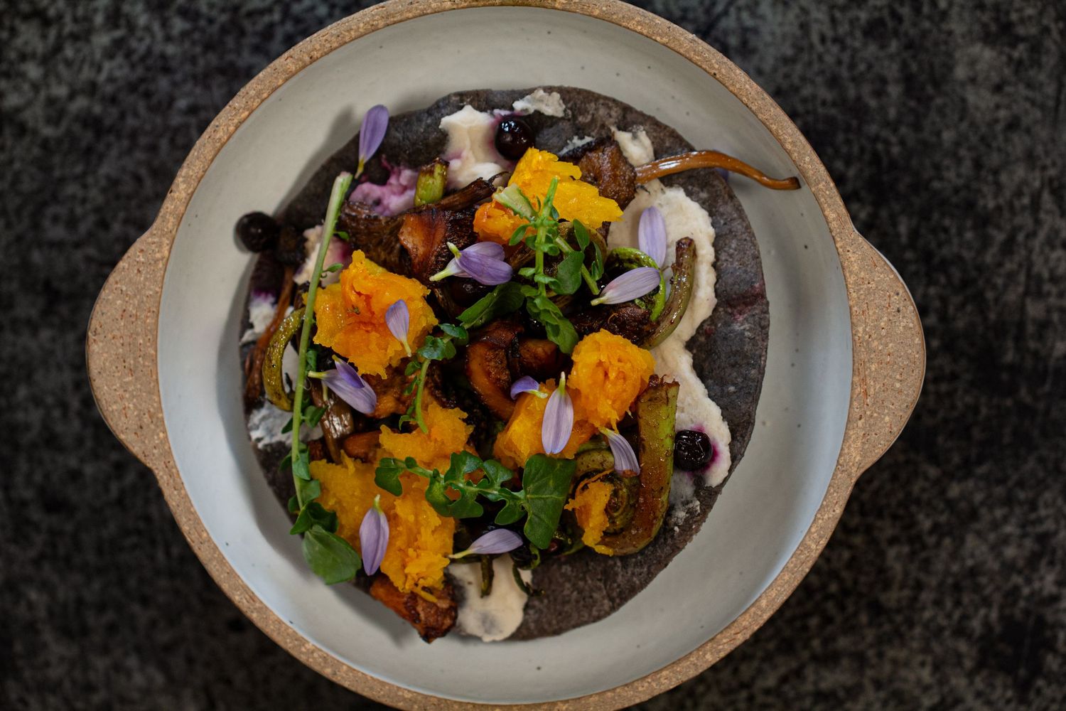 Blue corn tortilla topped with colorful mushrooms, squash cubes, and fresh herbs served in a rustic ceramic dish.