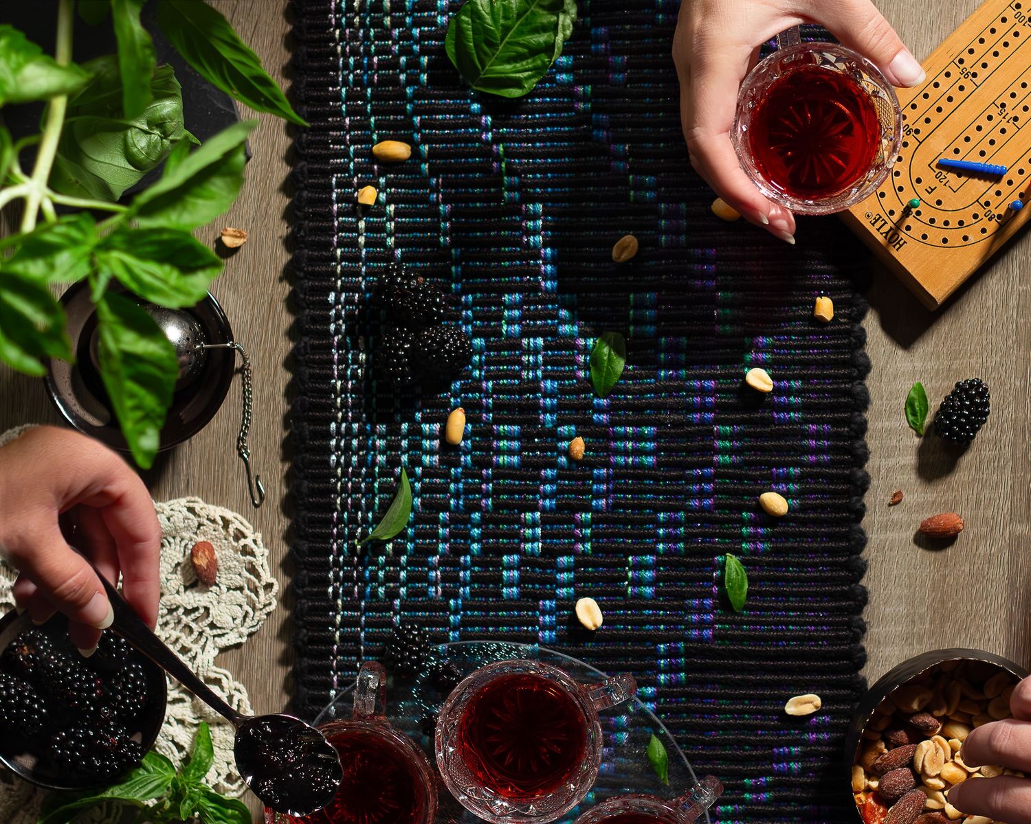 Hands pouring tea into glasses with cheese, nuts, and greenery on a dark rustic table setting.