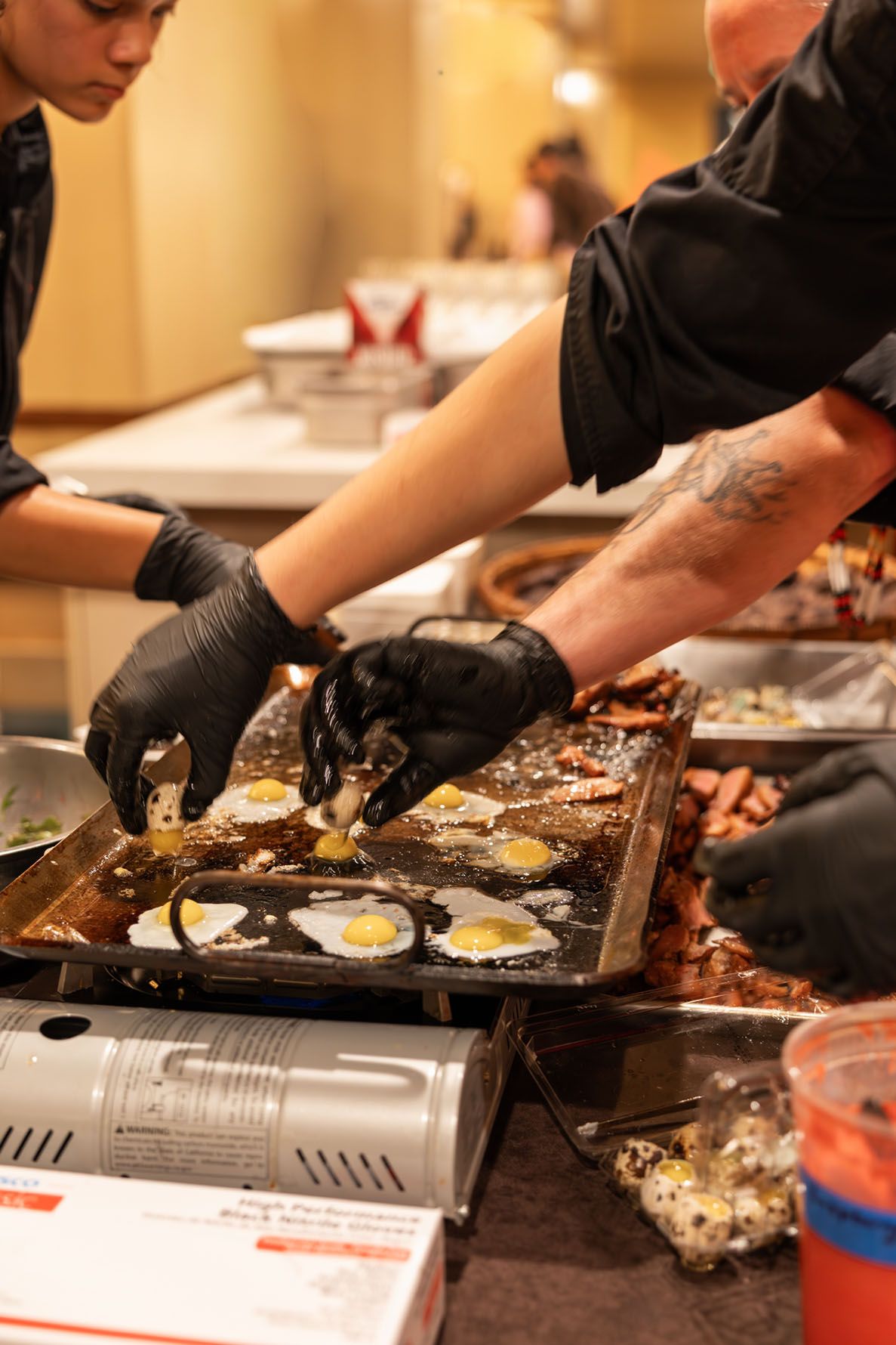 Chefs in black gloves cook quail eggs and other ingredients on a large commercial flat top griddle.