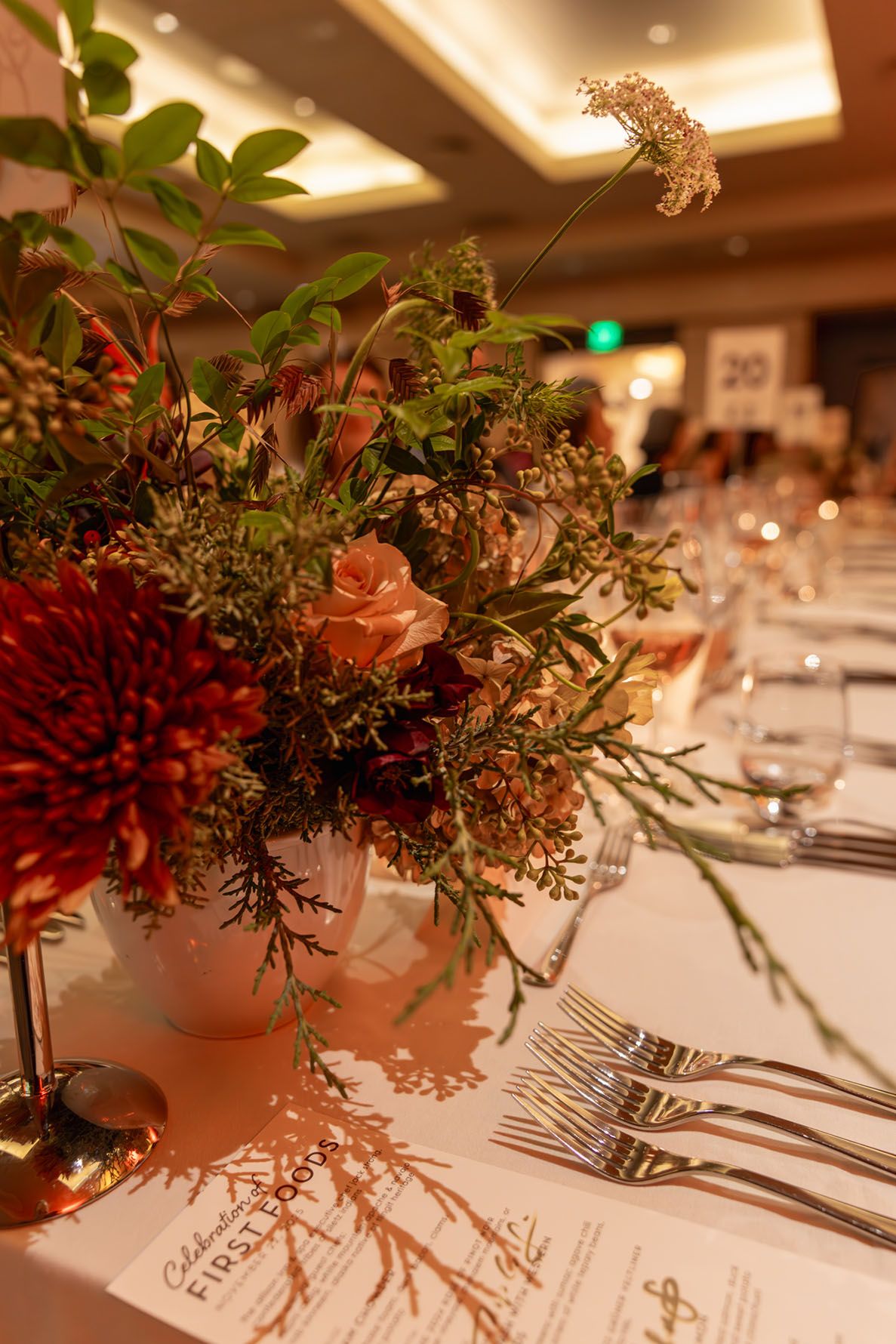 Elegant floral centerpieces with greenery and red blooms cast shadows on white tablecloths at a formal dining event.