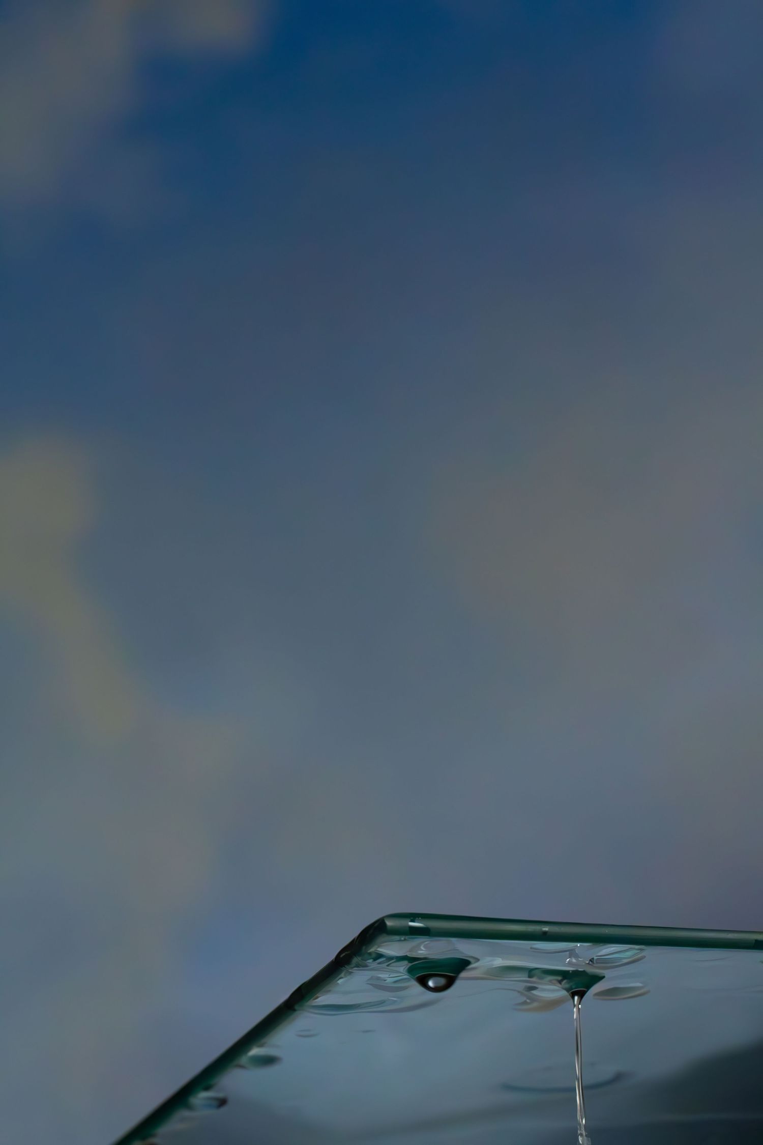 Raindrops fall on a dark car windshield against a moody blue sky creating a reflective pattern.