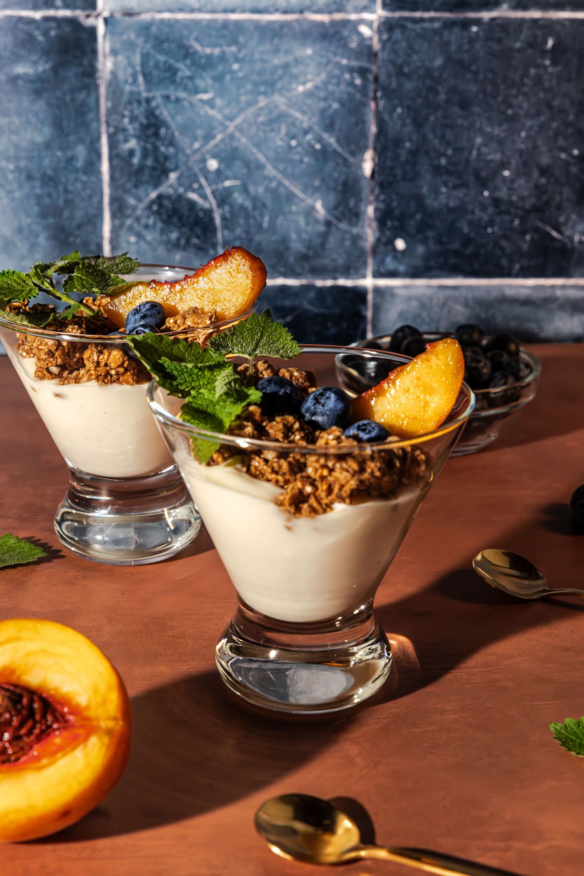 Yogurt parfait cups with granola, fresh fruit and mint garnish arranged in a row on a dark rustic surface and dark blue tile background.
