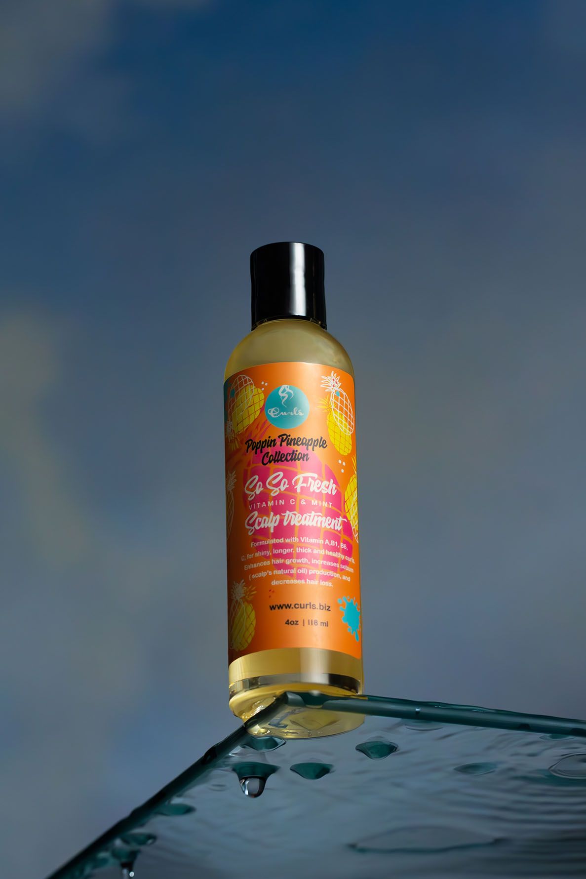 A bottle of Curls hair oil sits atop a glass surface against a moody blue sky background with water droplets on the glass.
