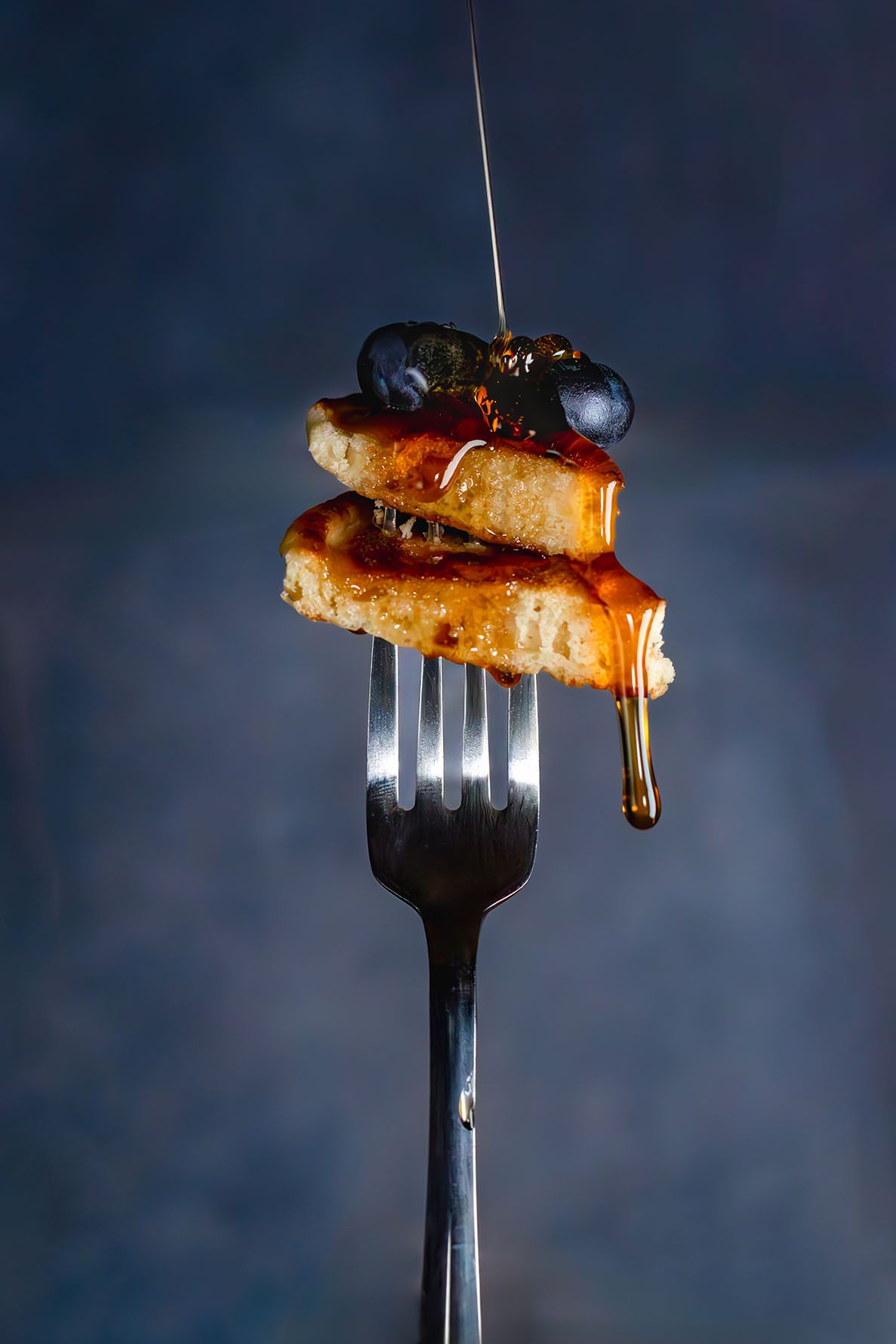 Two stacked pieces of pancakes with fresh blueberries balanced on a silver fork against a blue background and syrup being poured on them.
