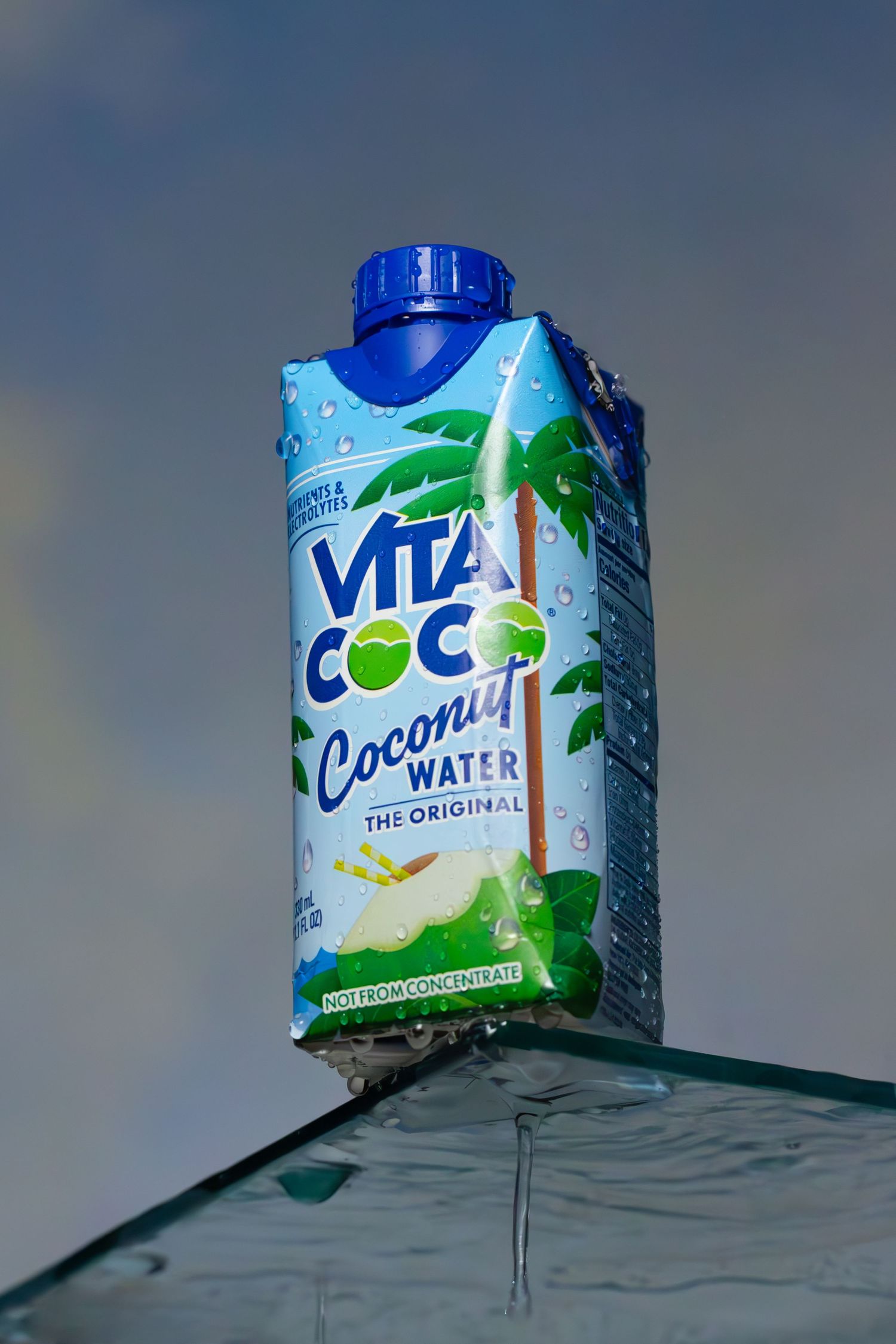 A carton of Vita Coco coconut water with palm tree graphics sitting on clear glass surface against blue sky background.