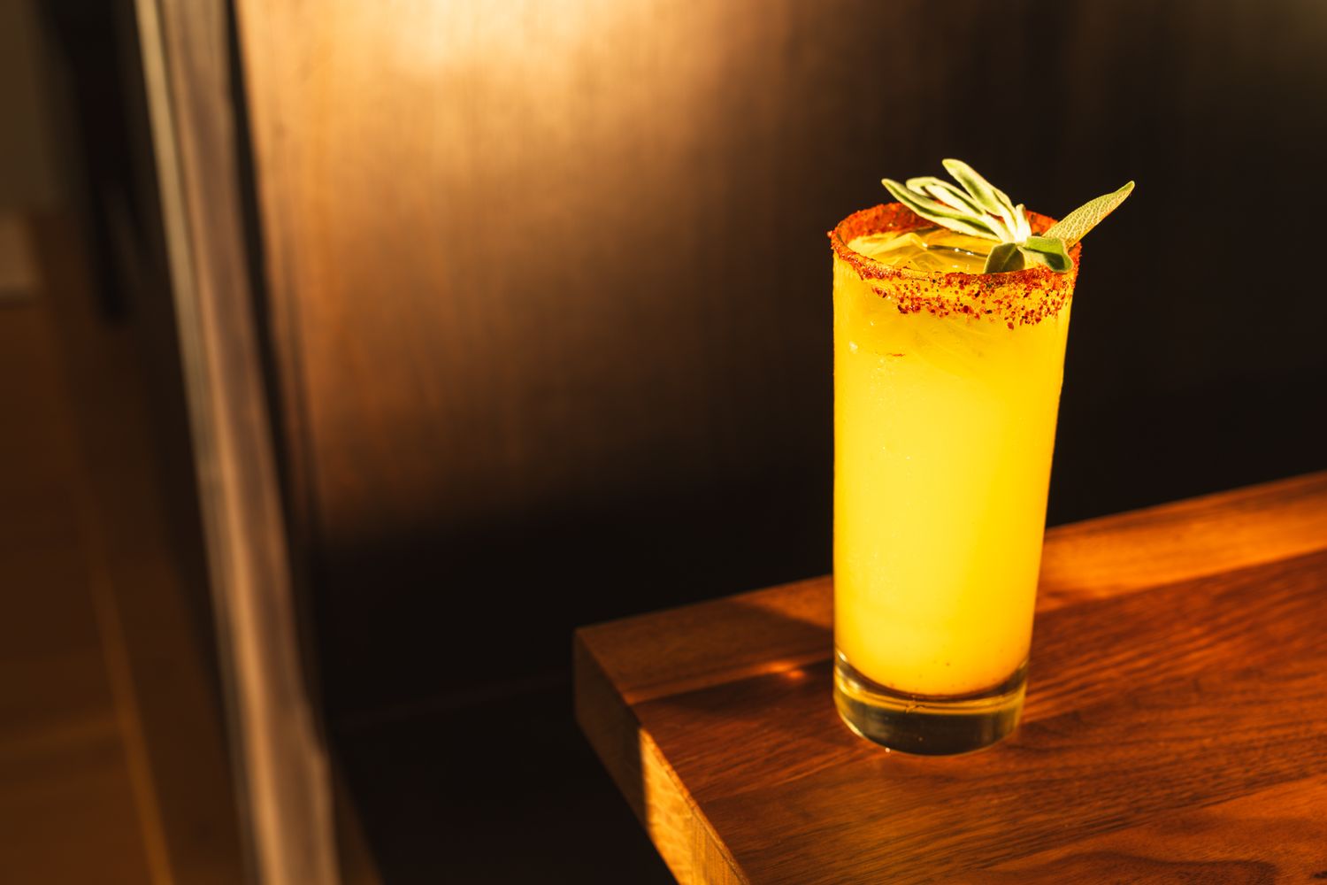 A vibrant yellow mocktail garnished with safe and chili salt rim in a tall glass on a wooden bar. B. Steele Images, beverage photography