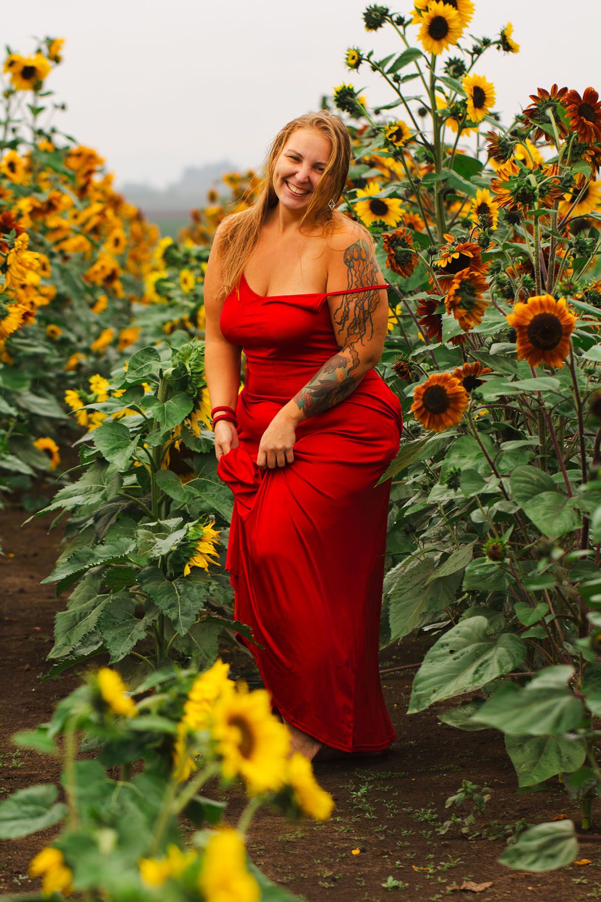 Brigit Steele is in a red dress that stands out beautifully against tall blooming sunflowers in a garden field in the early Fall