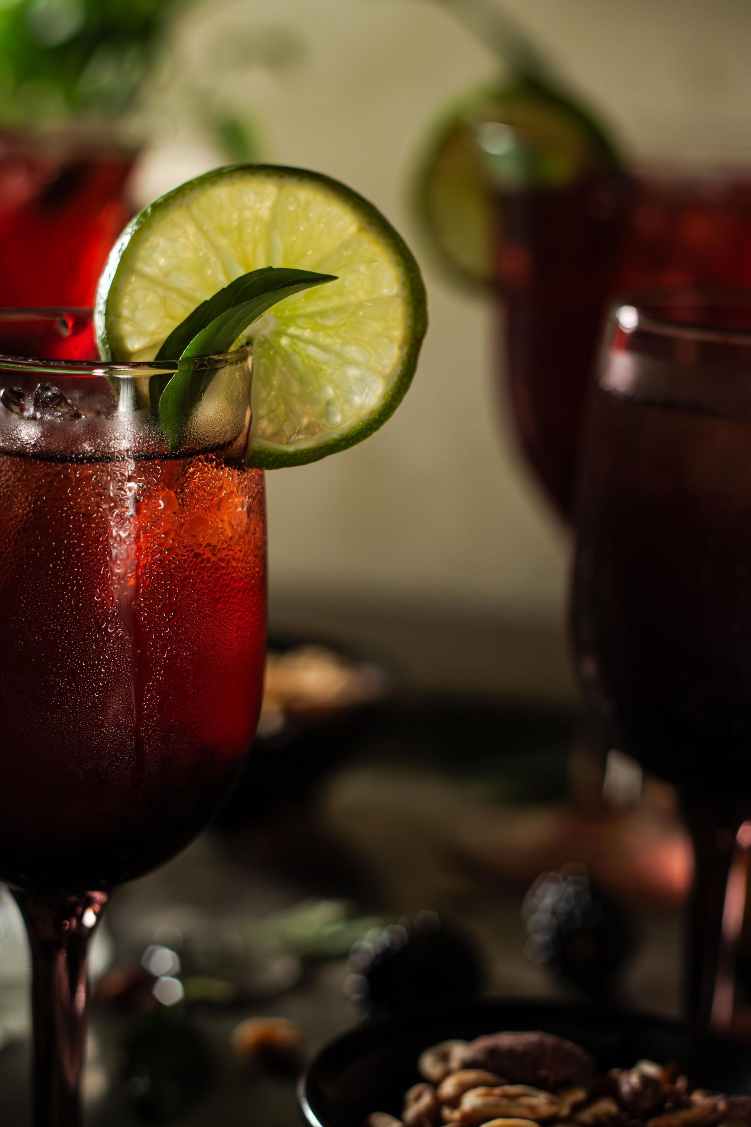 Dark red mocktail garnished with fresh lime slice and basil leaf in wine glass against moody background with scattered nuts.