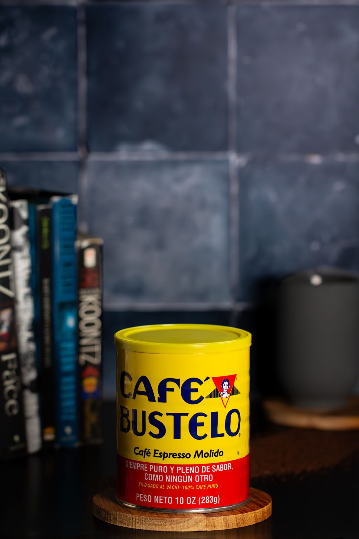 A yellow can of Cafe Bustelo espresso coffee sits on a dark surface with books in the background. Brigit Steele, photography services.