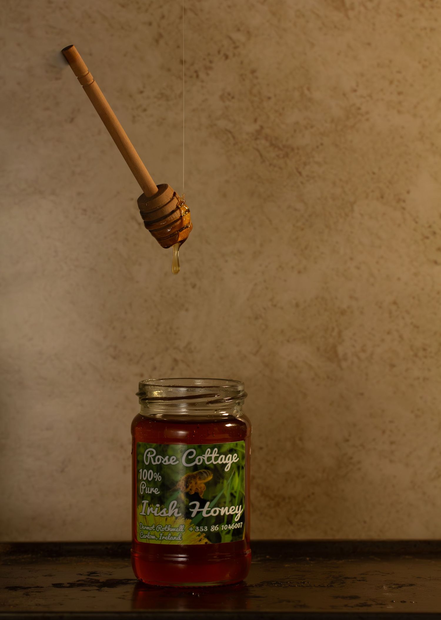 A wooden honey dipper drips honey over a glass jar of Rose Cottage fresh honey on a rustic wooden surface.
