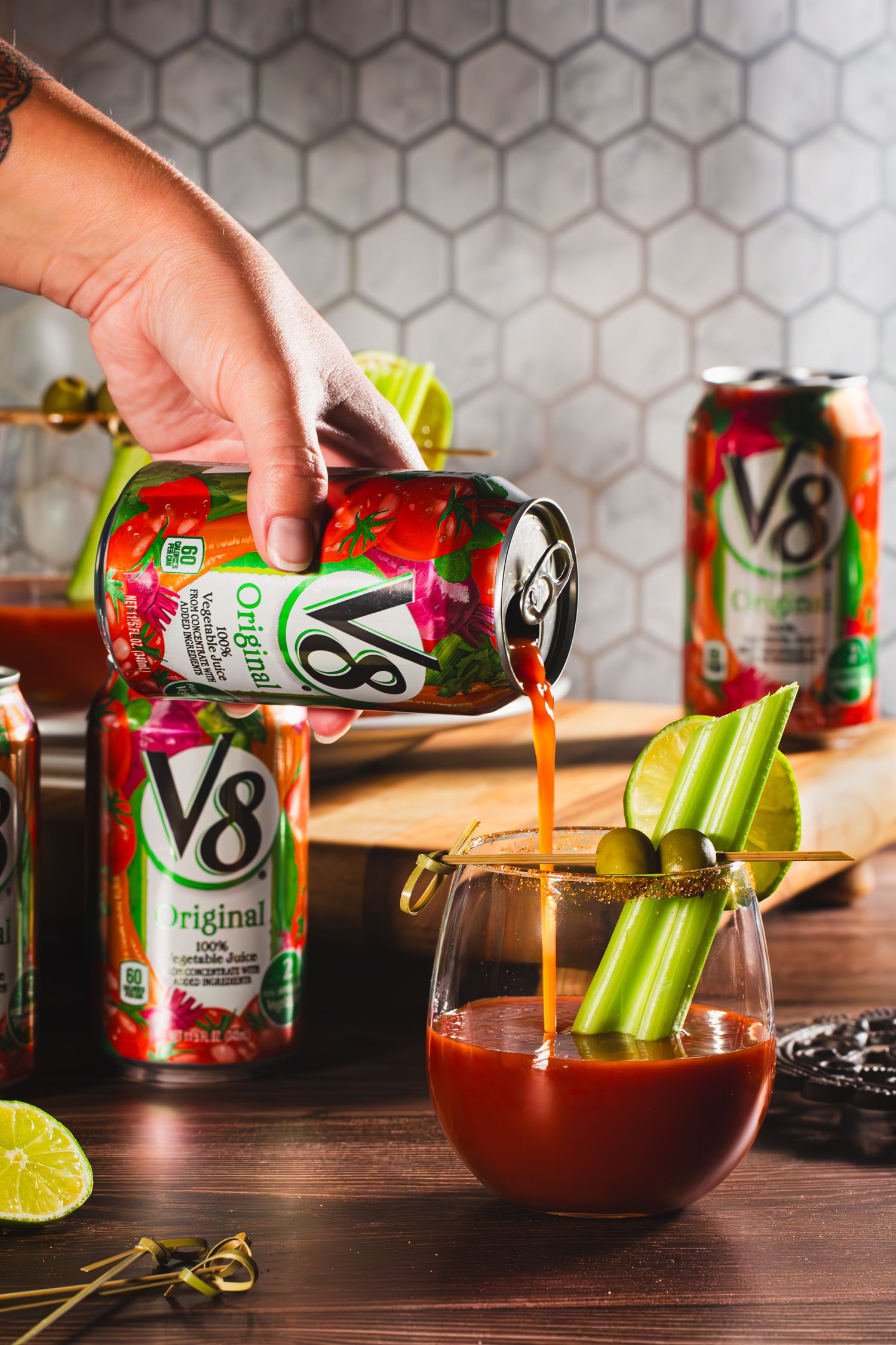 V8 vegetable juice being poured into a mocktail glass garnished with celery sticks and lime on a wooden table.