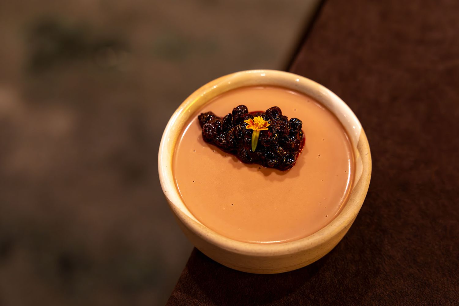 A creamy pinenute dessert served in a white ceramic bowl topped with dark huckleberry-sage compote and edible flower garnish.