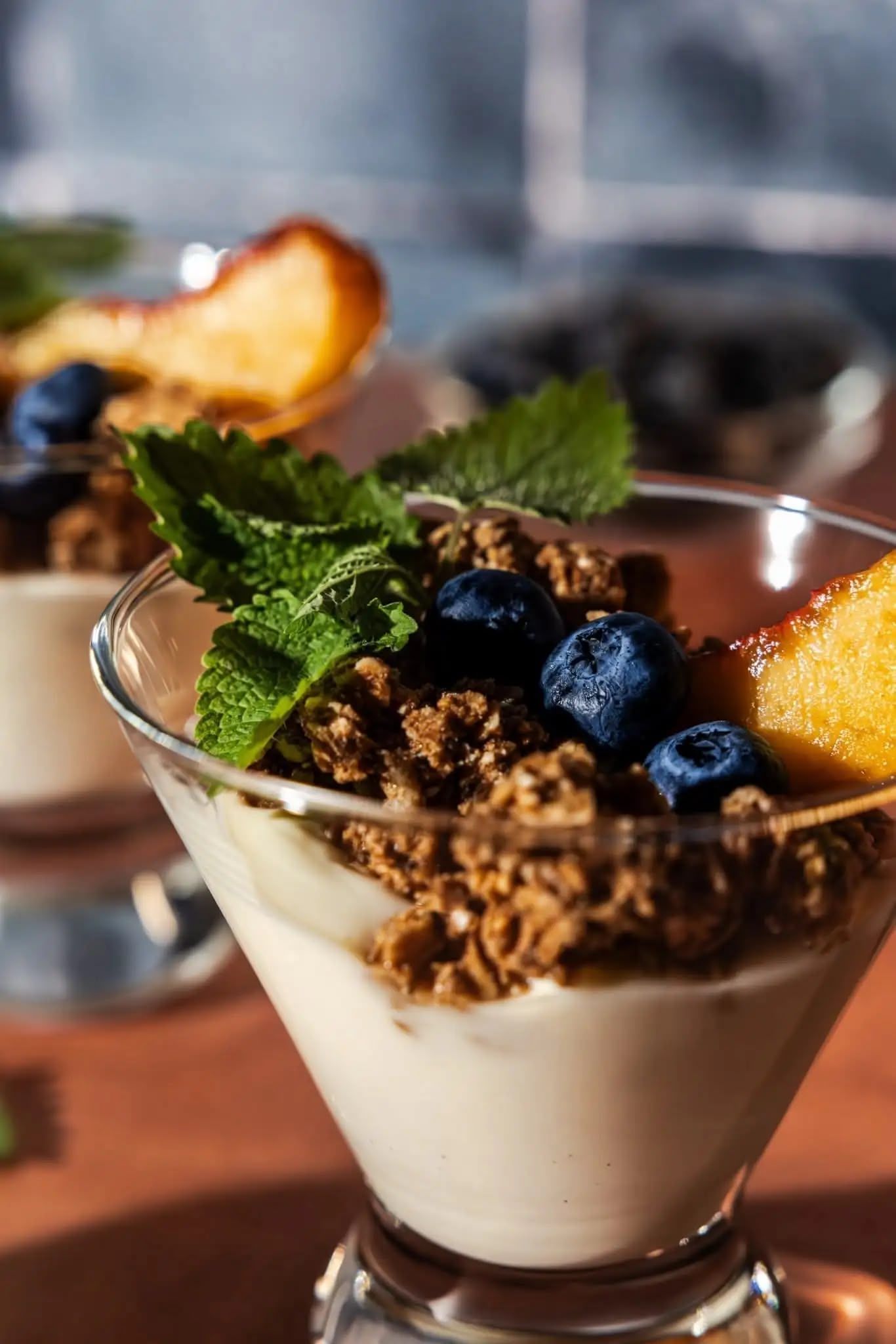 Peach Parfait with creamy white base, granola crumbles, fresh blueberries, and mint leaves served in a glass. Food and beverage photography