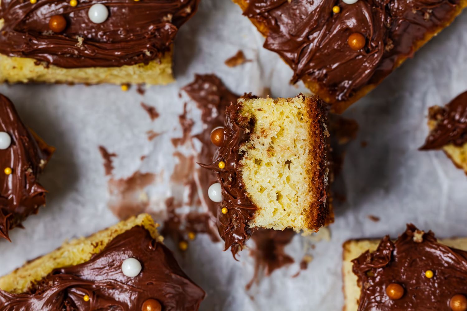 Yellow cake with chocolate frosting and colorful candied balls on the cake pieces arranged in a rustic display on parchment paper. B. Steele