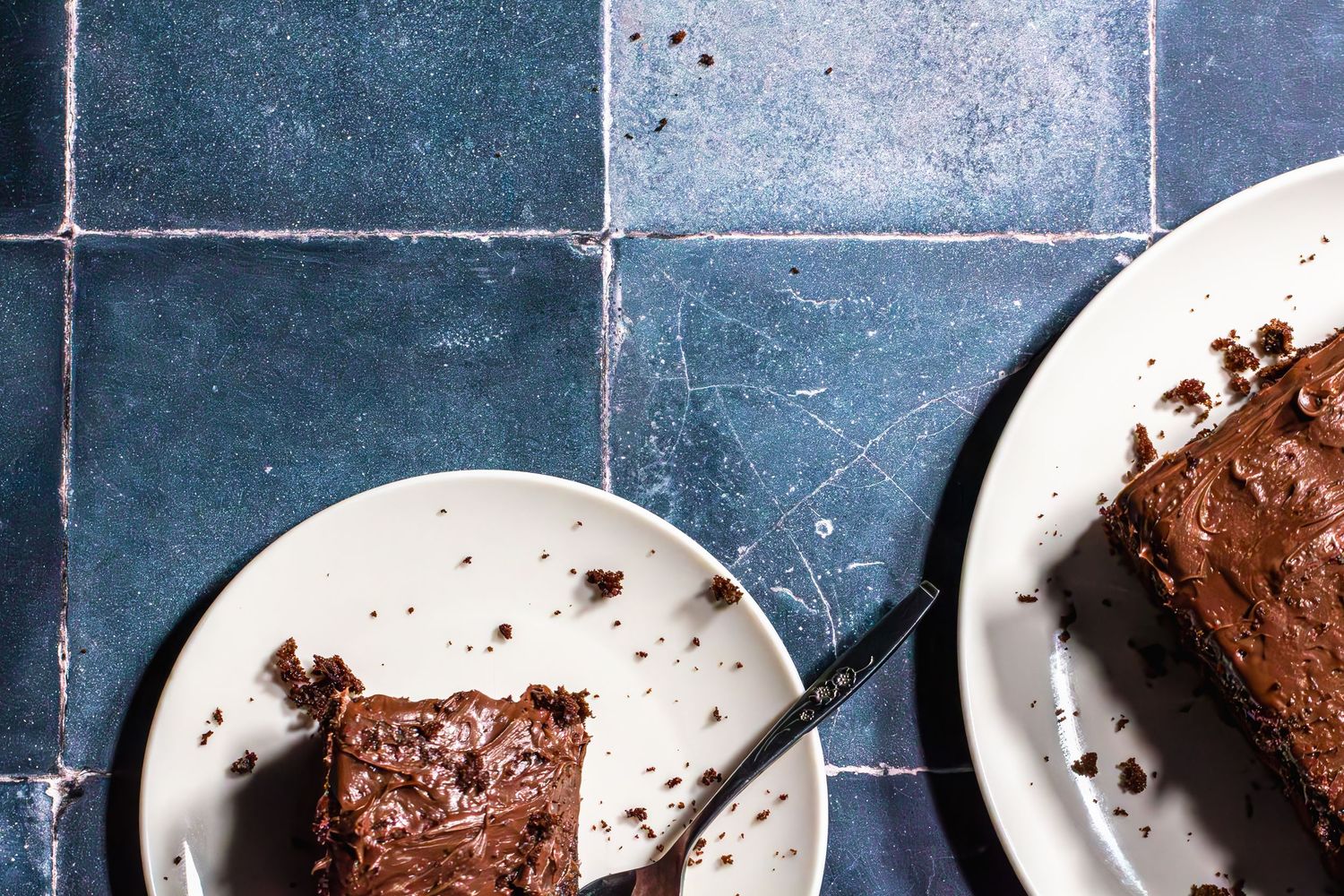 Two plates of rich chocolate cake slices with fudgy frosting on a blue tiled surface, one slice partially eaten.