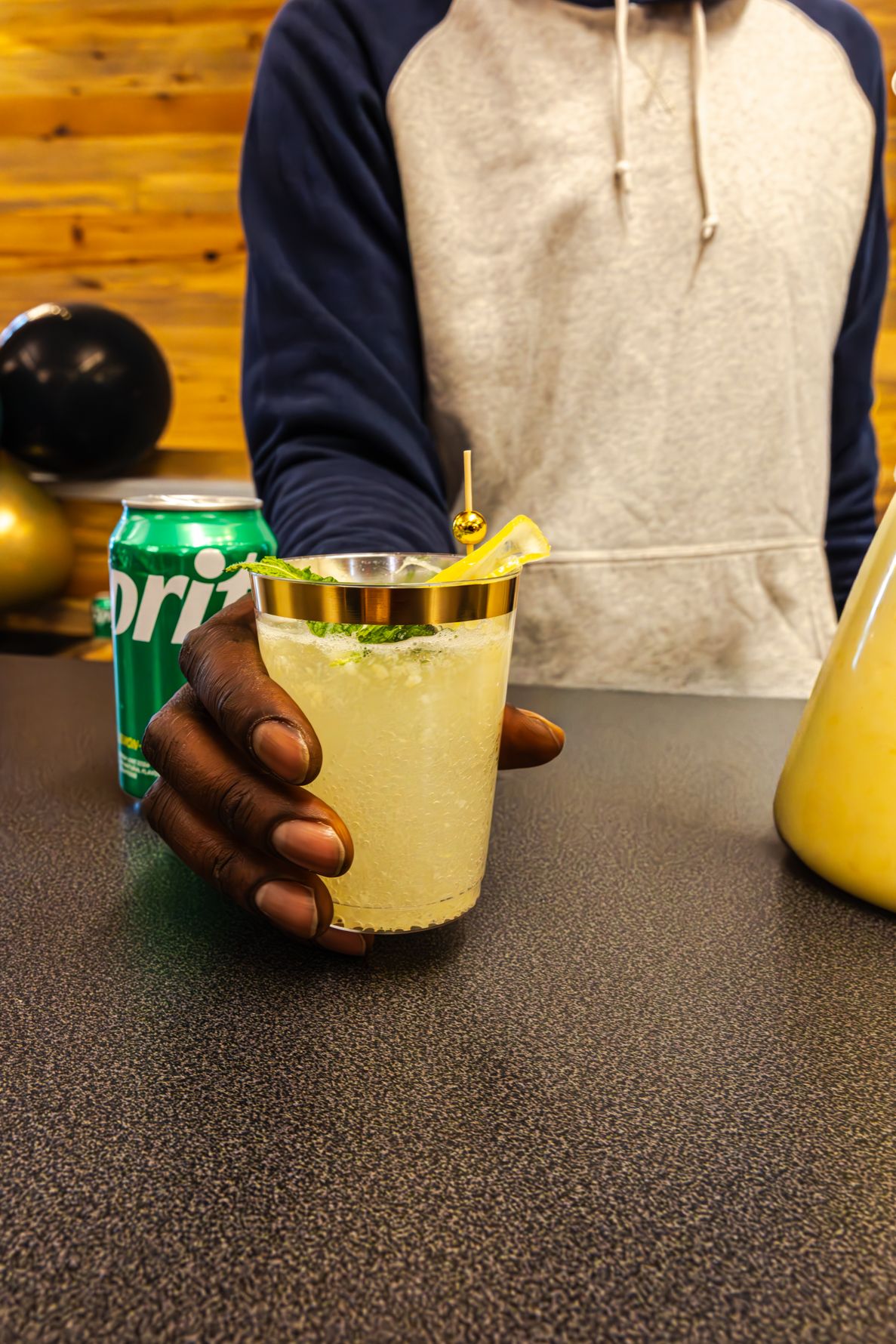 Person holding a frosted cocktail glass with mint garnish beside a Sprite can on a bar counter. B. Steele Images. Food & Beverage photograph