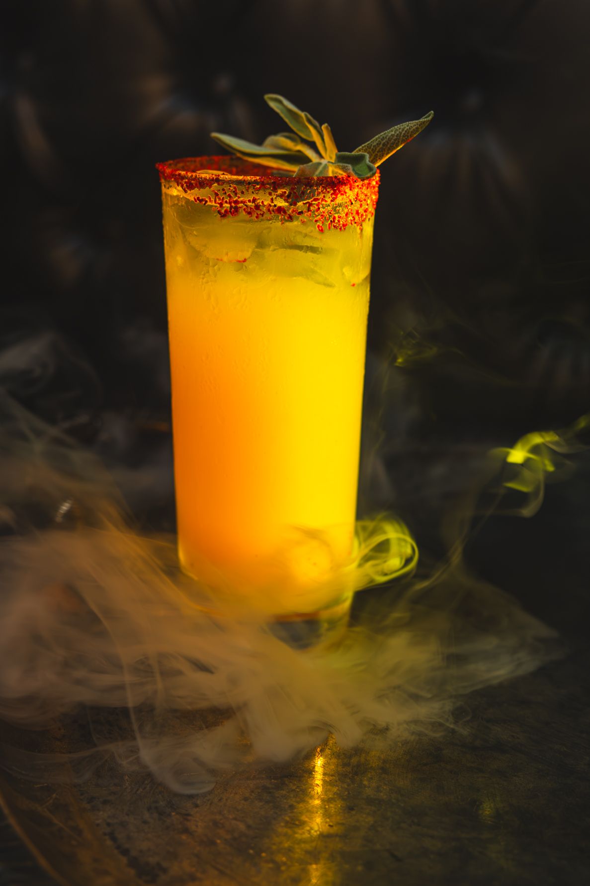 A vibrant yellow mocktail garnished with safe and chili salt rim in a tall glass on a dark sofa. B. Steele Images, beverage photography