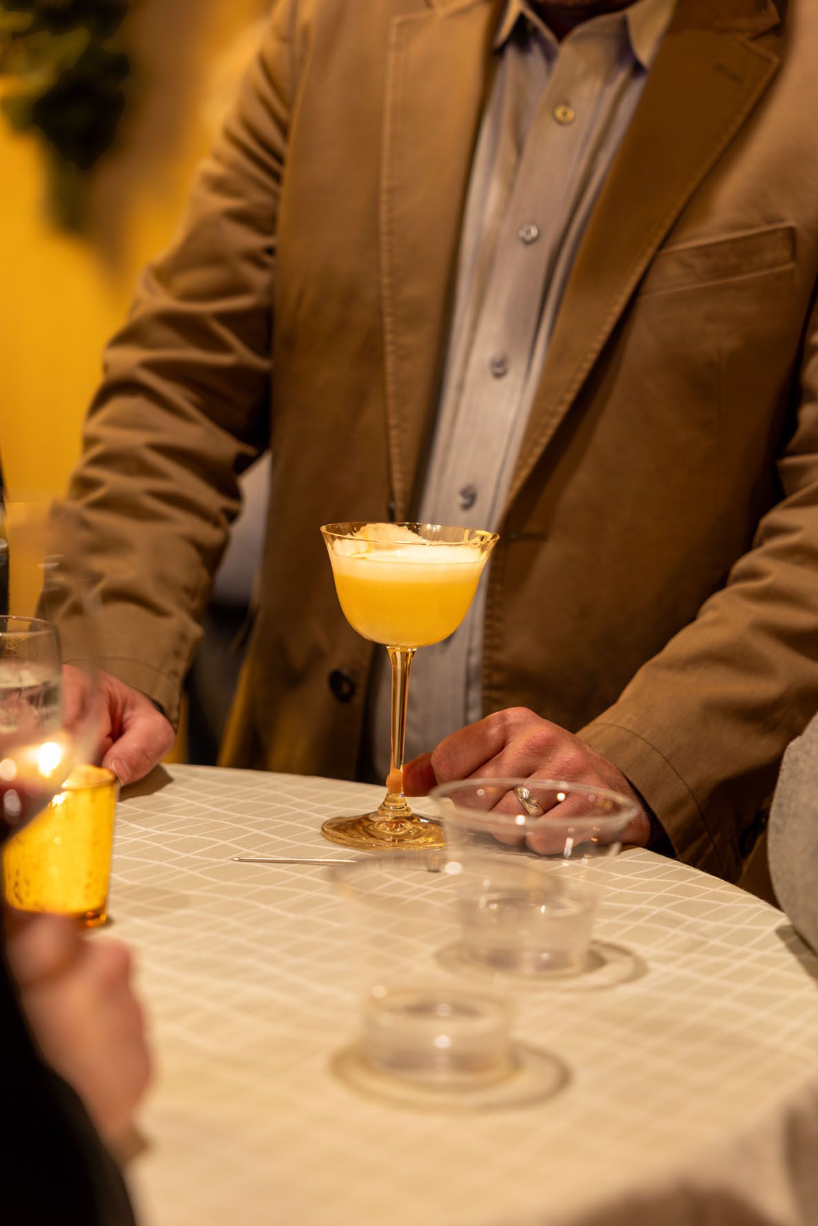 A yellow cocktail in a coupe glass sits on a white tablecloth as someone in a brown blazer stands at high-top table.