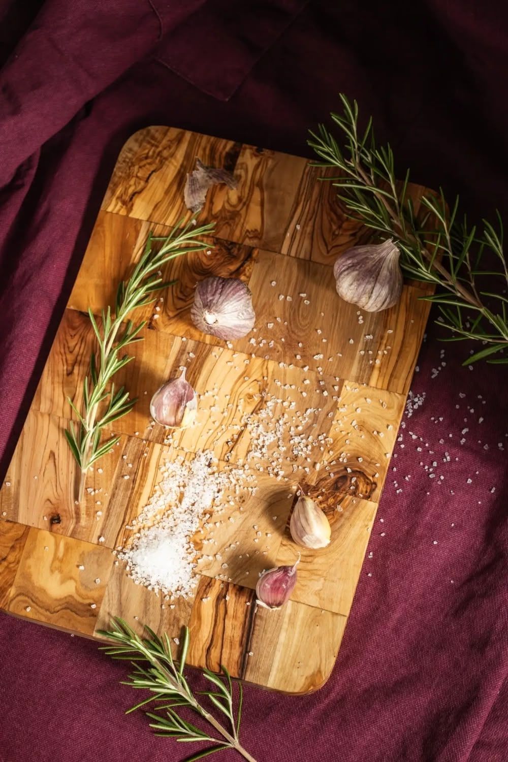 Wooden cutting board with fresh rosemary sprigs, garlic cloves, and scattered sea salt on a burgundy tablecloth. Food styling, Brigit Steele