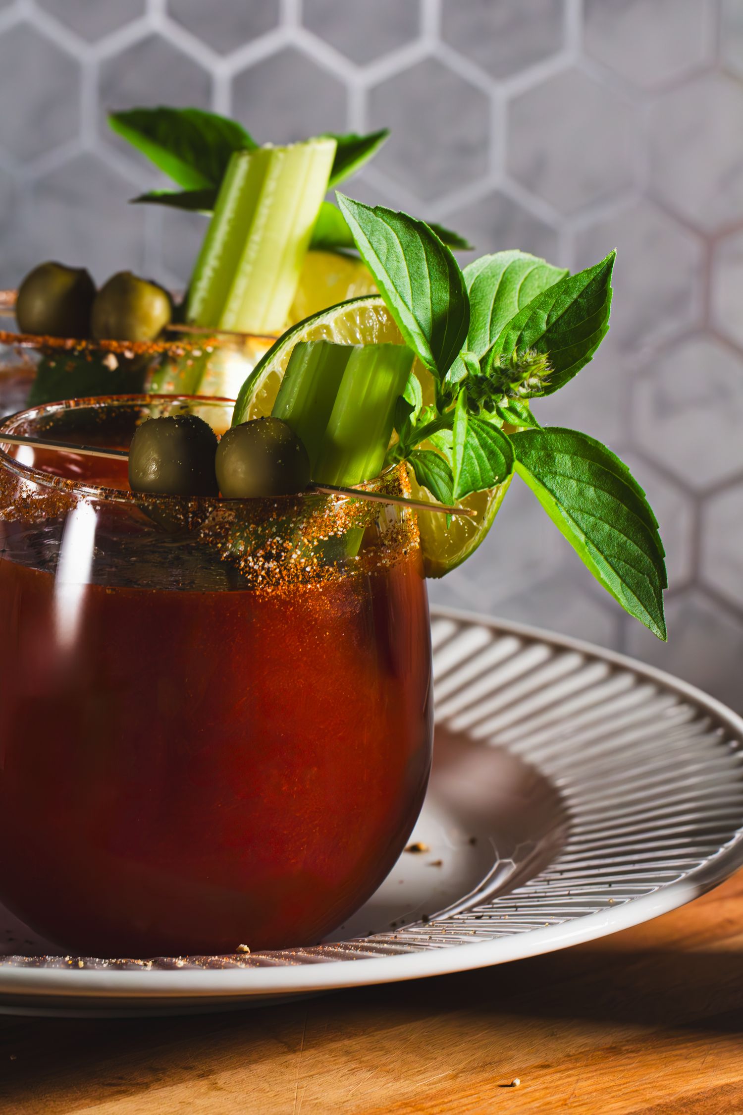 A garnished Bloody Mary mocktail with fresh basil, celery stalk, and olives sits on a ridged plate against a tiled background on wood table.