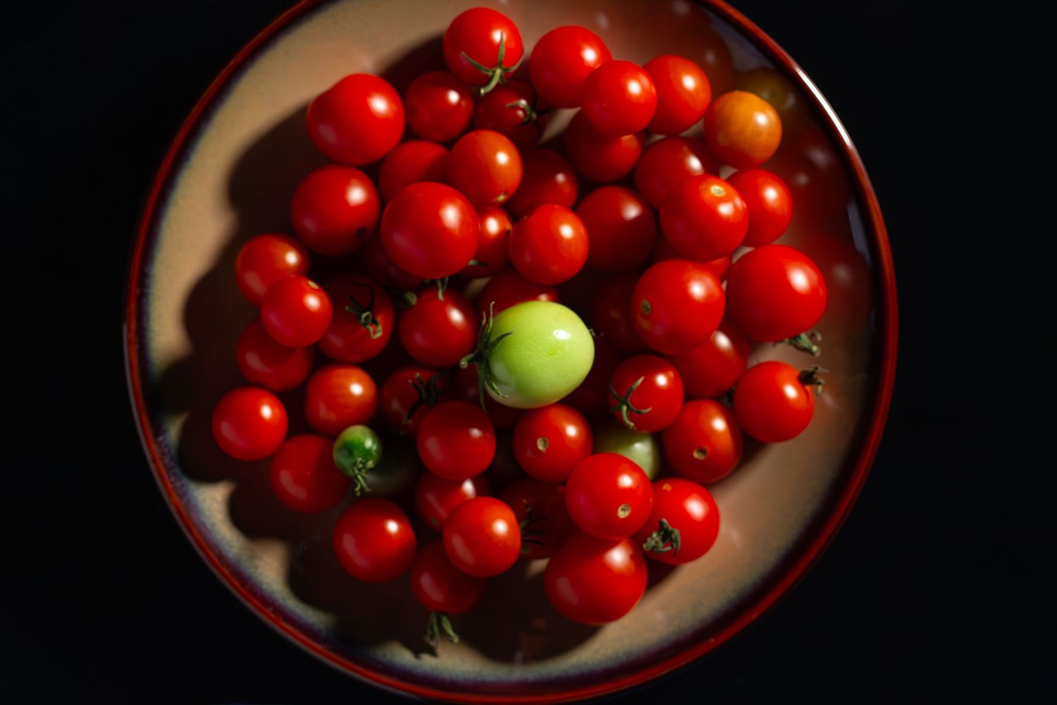 A plate of bright red cherry tomatoes with one green tomato standing out among them on a black background. B. Steele Images, food photograph