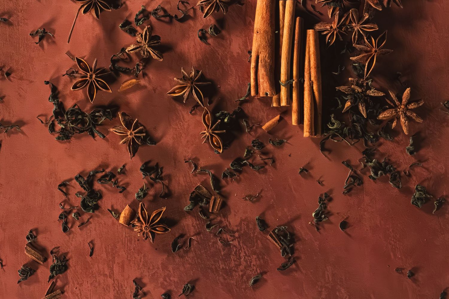Star anise, cinnamon sticks, and dried herbs scattered on a rich red-brown surface, perfect for warm spice blends.