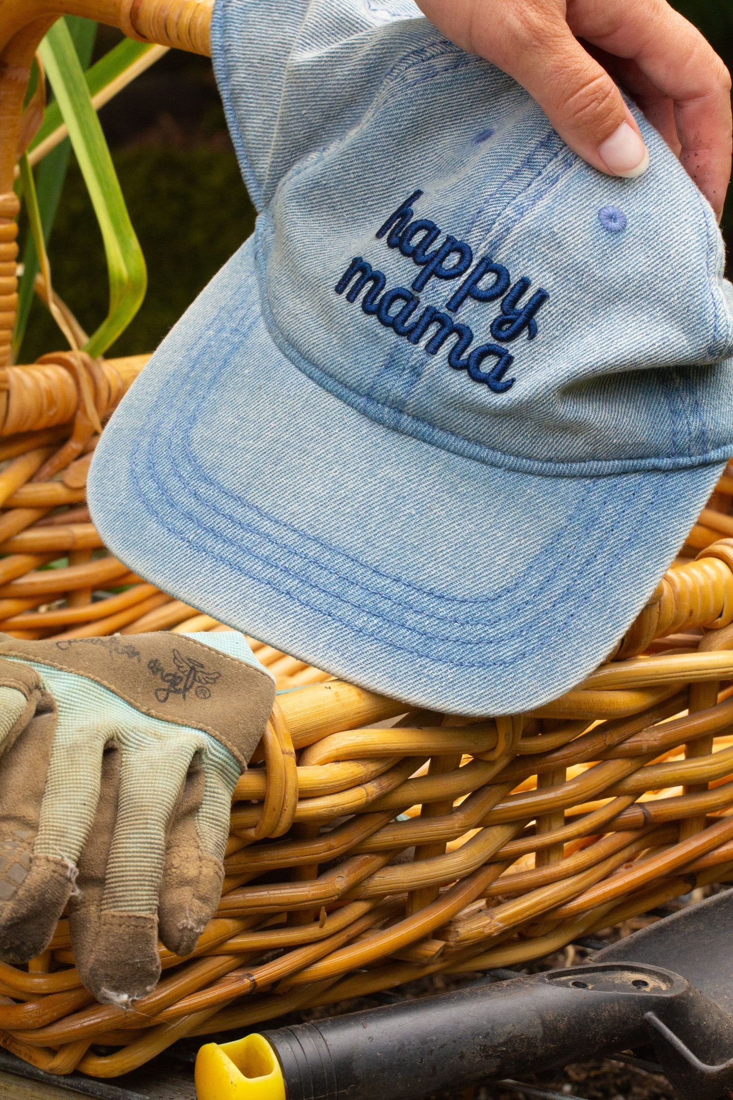 Light blue denim baseball cap with 'happy mama' text embroidered on it sits with gardening glove and tools with a wicker basket.