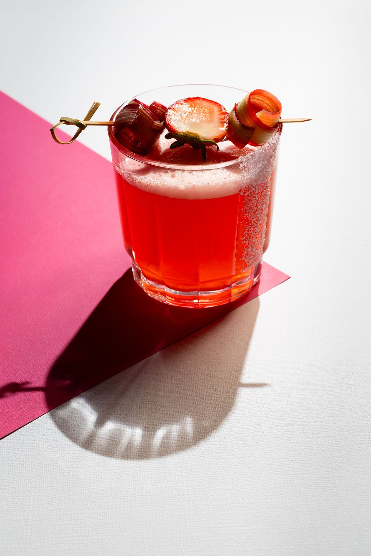 Non-alcoholic sugared rimmed strawberry rhubarb mocktail with fresh fruit garnish on white and pink backdrop. B. Steele Images
